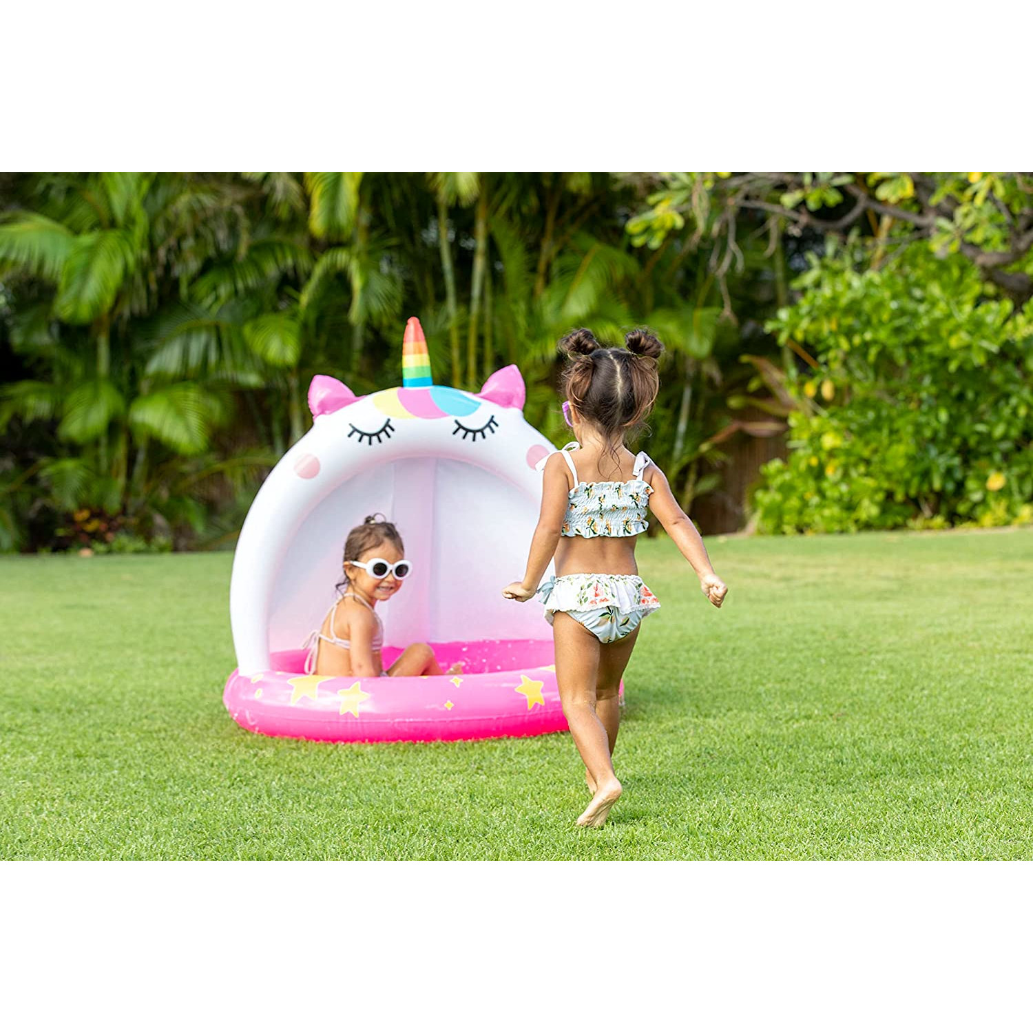 Zwei junge Mädchen in einem pinkfarbenen Einhorn-Pool. Ein Mädchen ist im Pool und das andere geht darauf zu. Grünes Gras.