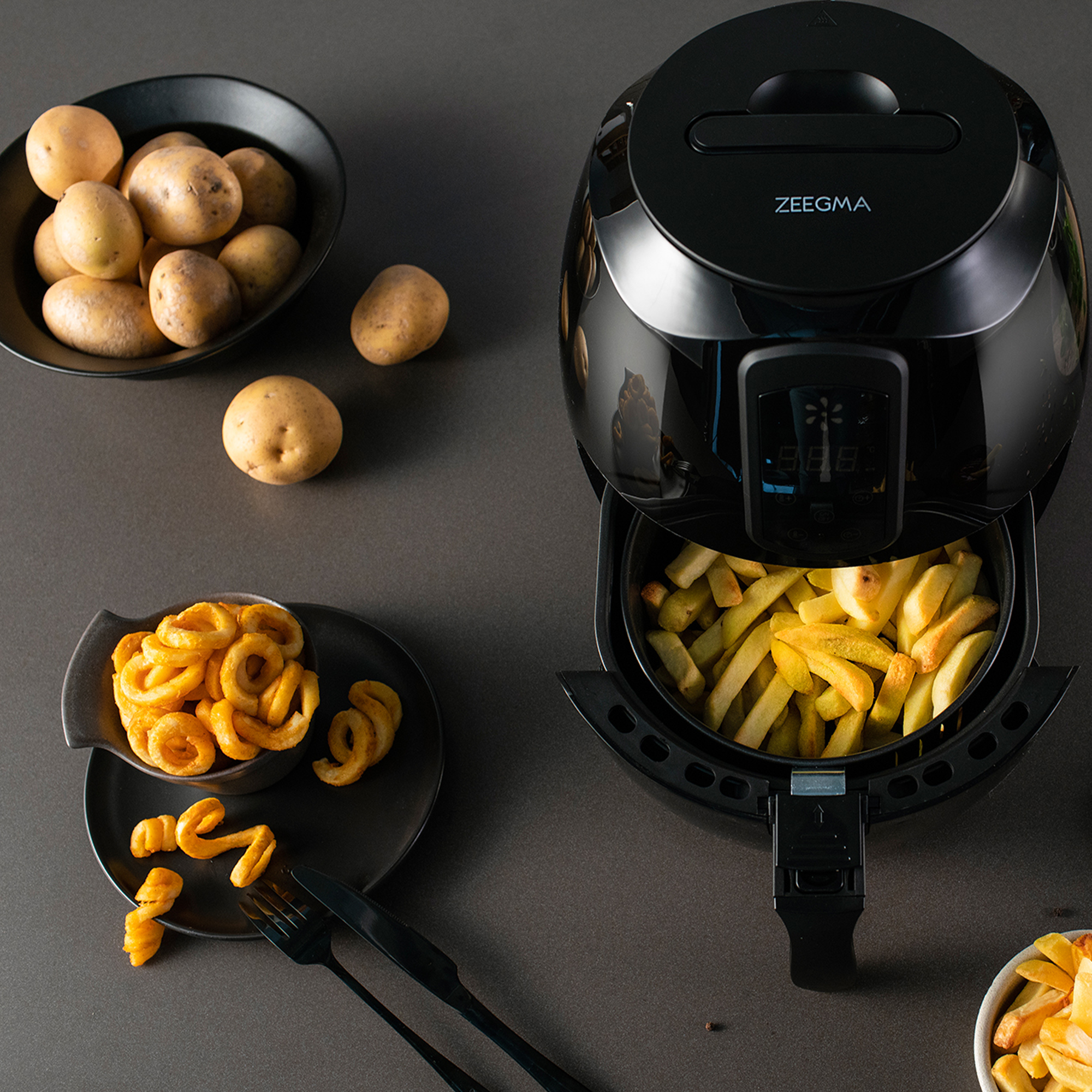 Czarna frytkownica beztłuszczowa, pełna frytek, i talerze z ziemniakami i frytkami kręconymi.