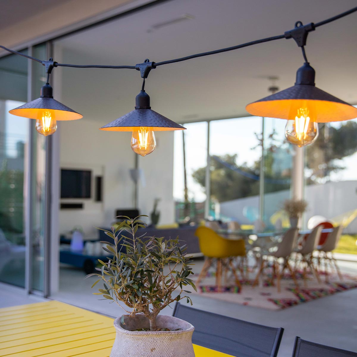 Terrasse im Freien mit Lichterketten. Gelber Tisch, kleine Pflanze und Blick auf einen Essbereich im Hintergrund.