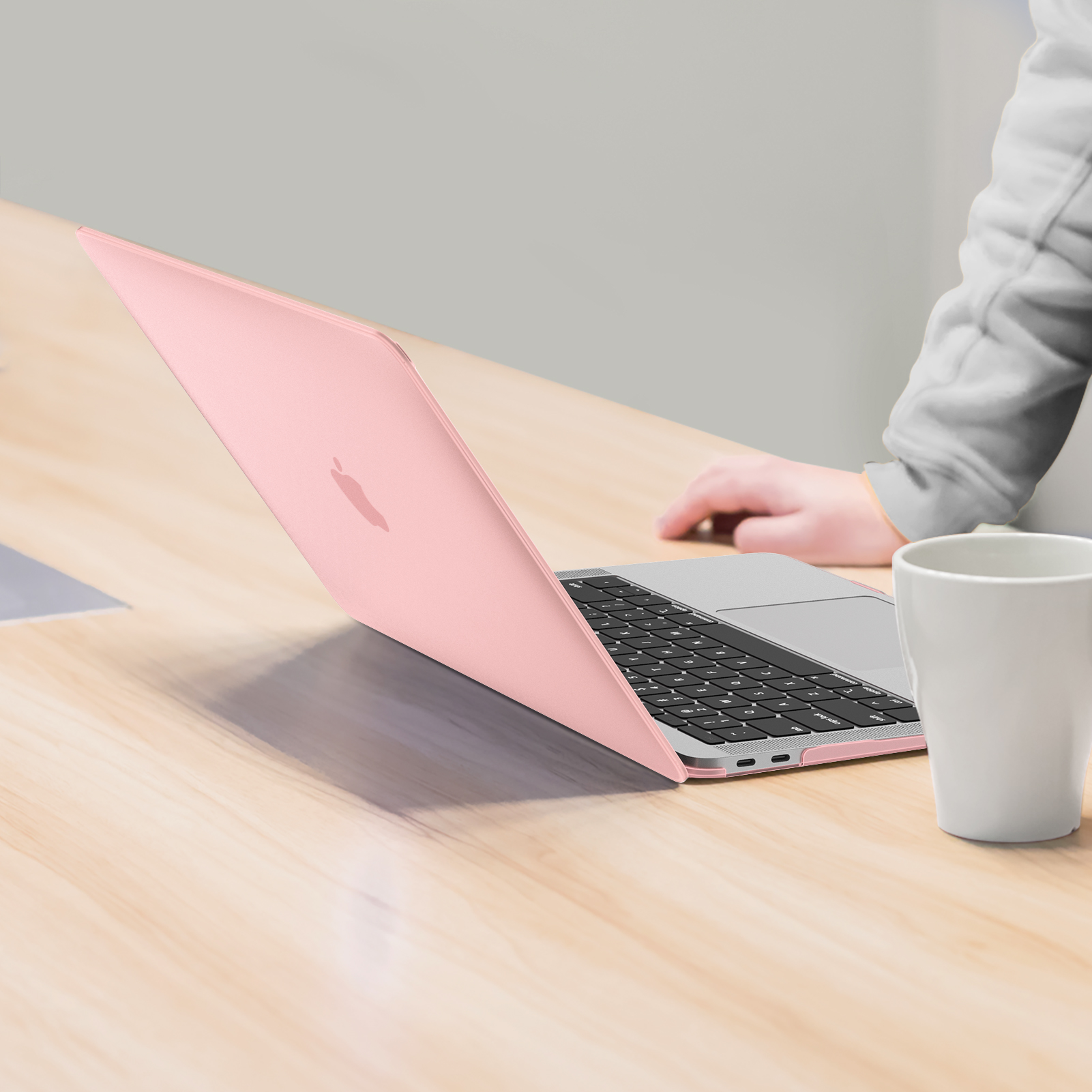 Ein pinkfarbener Laptop auf einem Holztisch. Eine weiße Tasse steht daneben, und die Hand einer Person ist zu sehen.