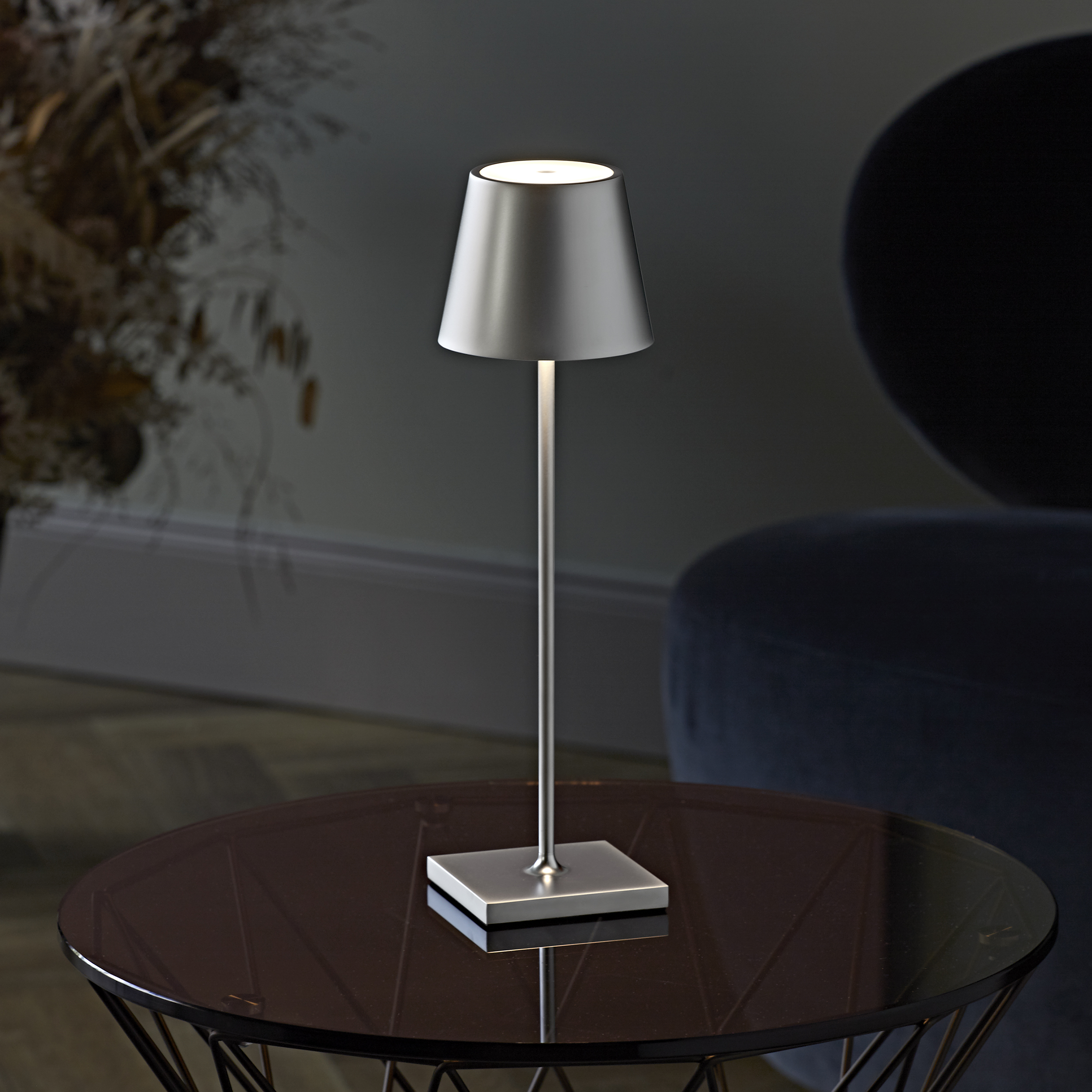 Lampe argentée sur une table. Pièce sombre avec vase et chaise bleue.
