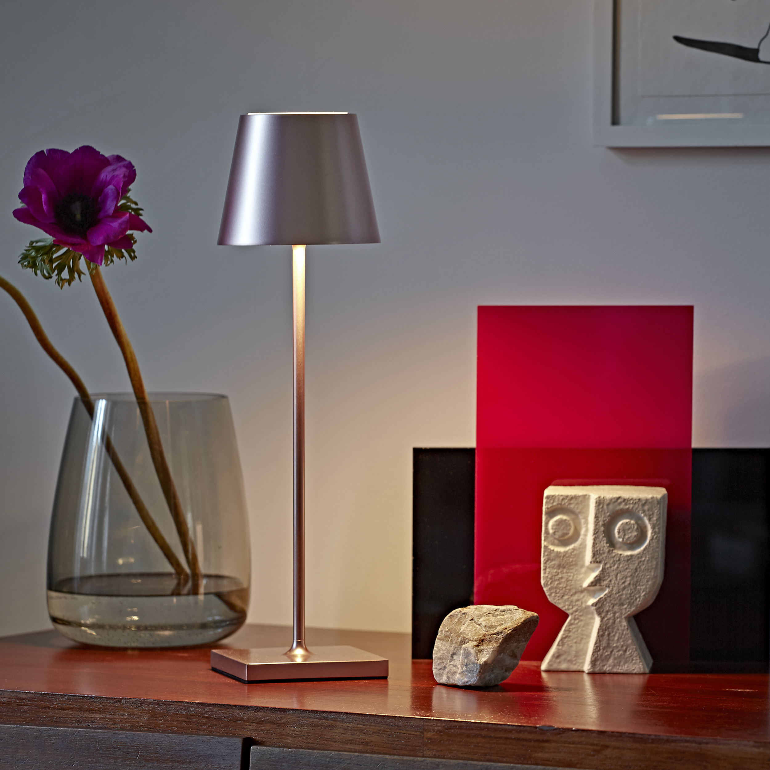 Eine beleuchtete Lampe mit Schirm, eine Vase mit Blumen, eine Skulptur und ein roter Block.