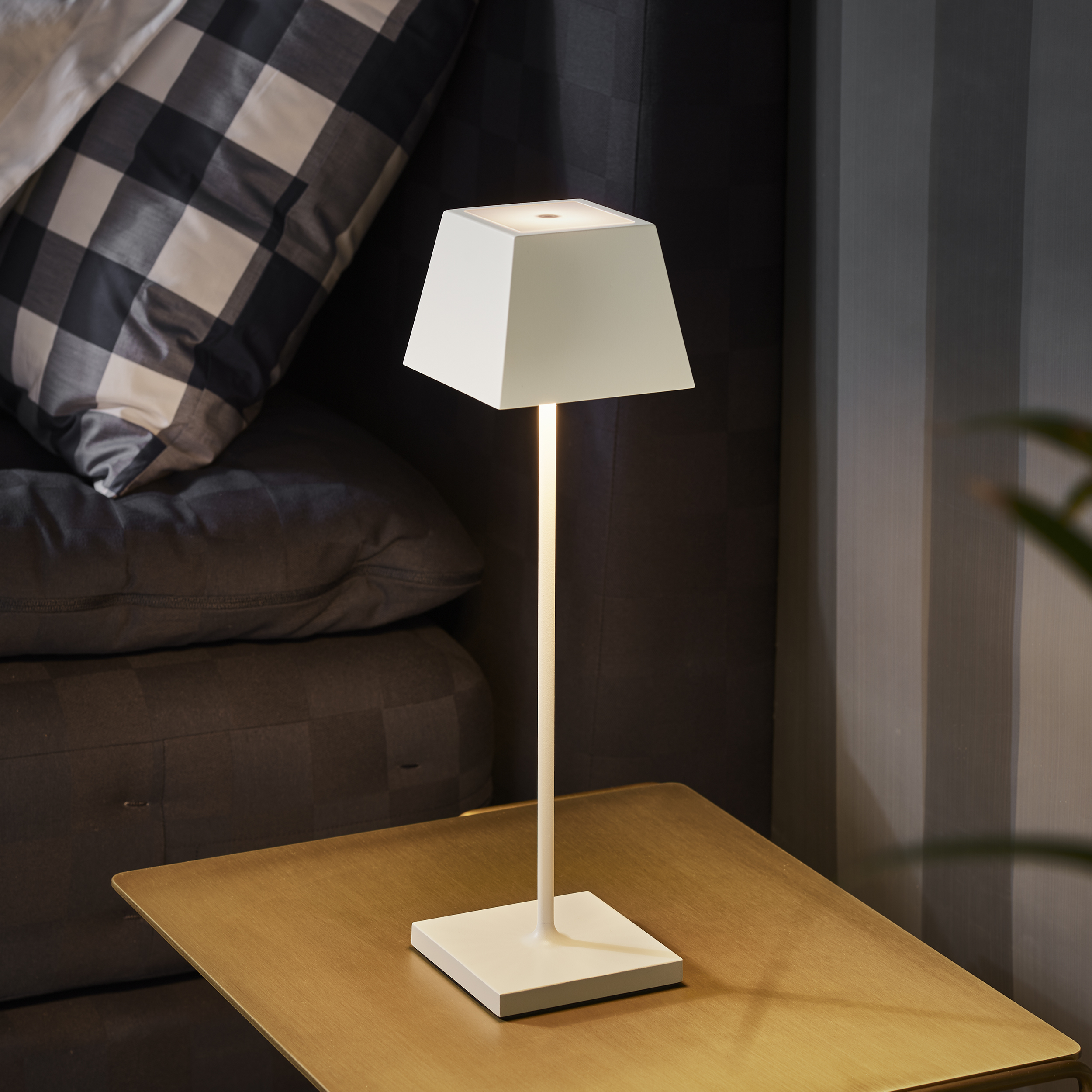 Eine weiße Lampe auf einem goldenen Tisch. Im Hintergrund sind ein Sofa und ein Kissen. Die Lampe ist an und strahlt Licht aus.