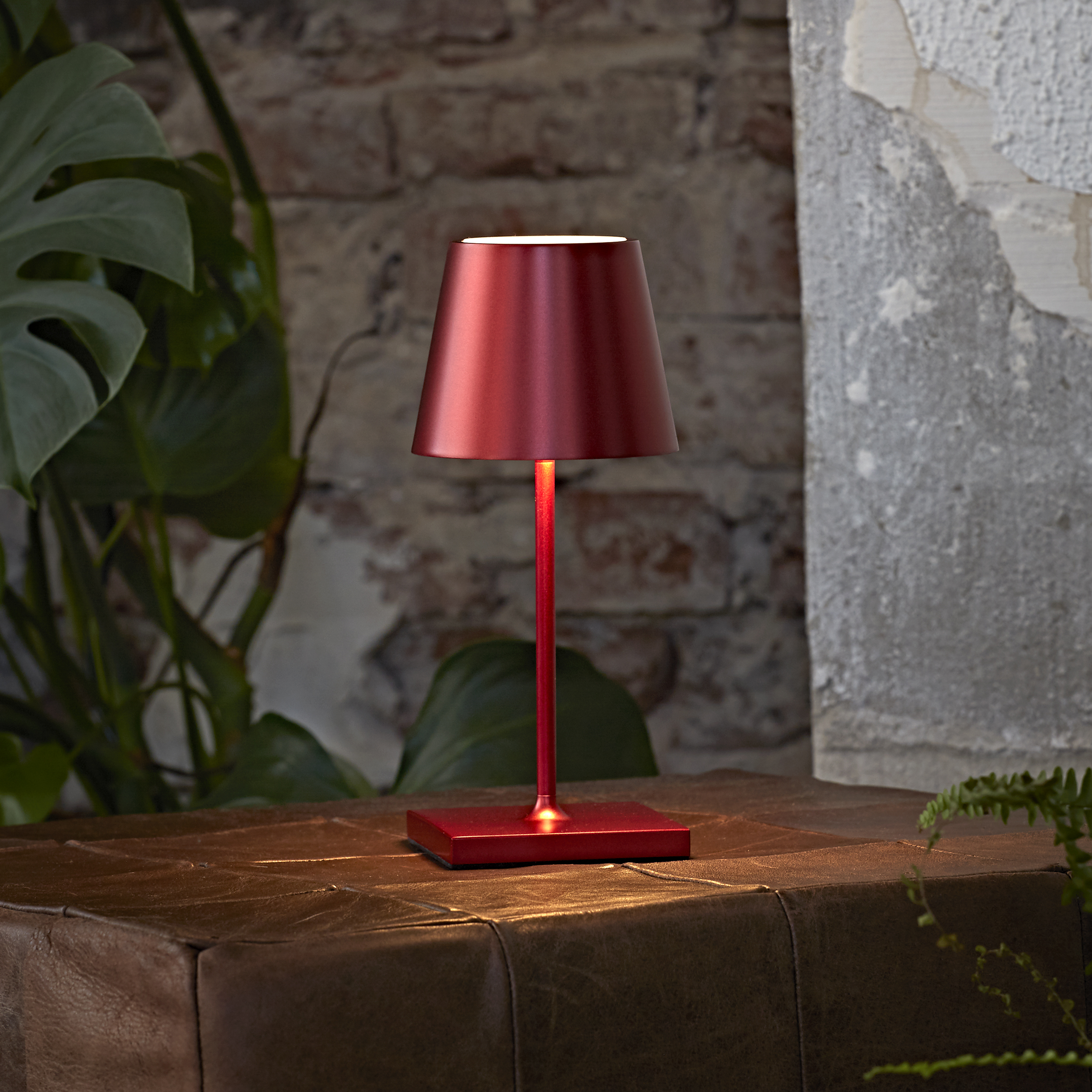 Eine rote Tischlampe mit konischem Lampenschirm auf einer braunen Lederoberfläche vor einer Backsteinmauer.