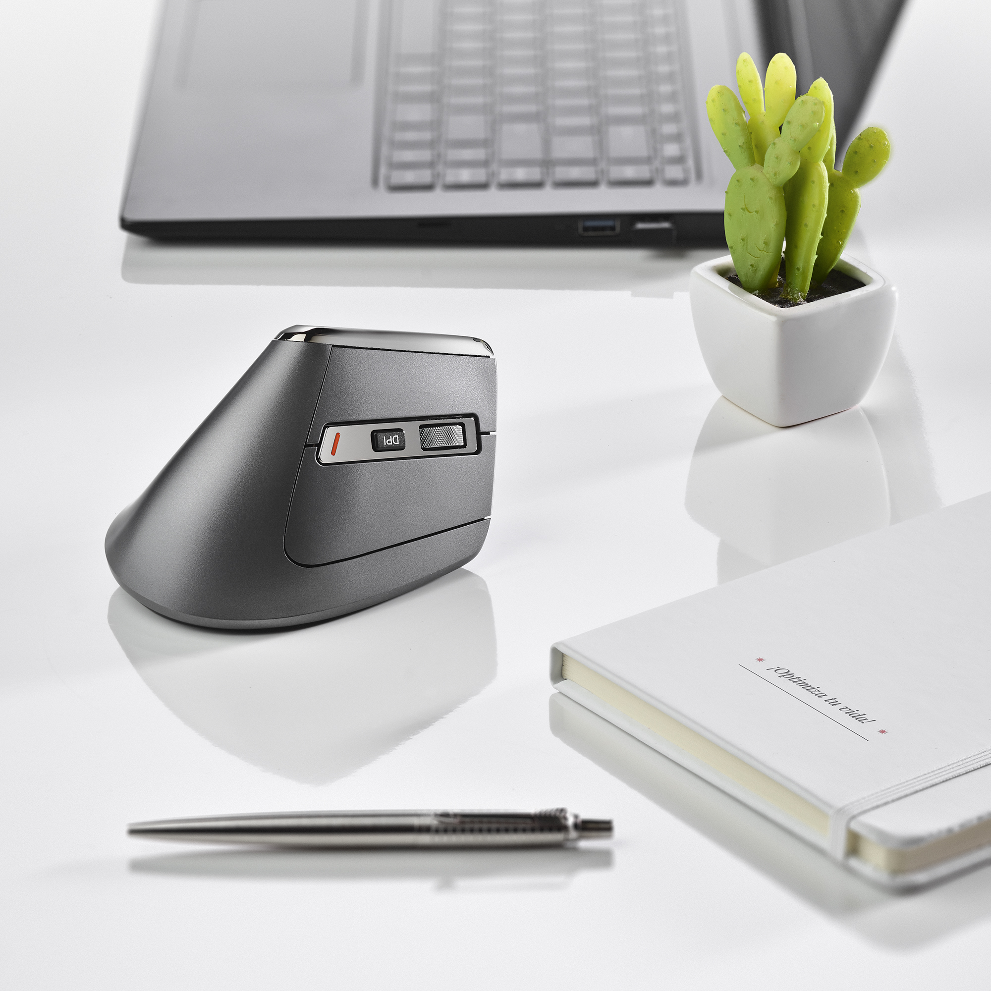 Mysz ergonomiczna, notatnik, długopis i mały kaktus na białej powierzchni. Laptop w tle.