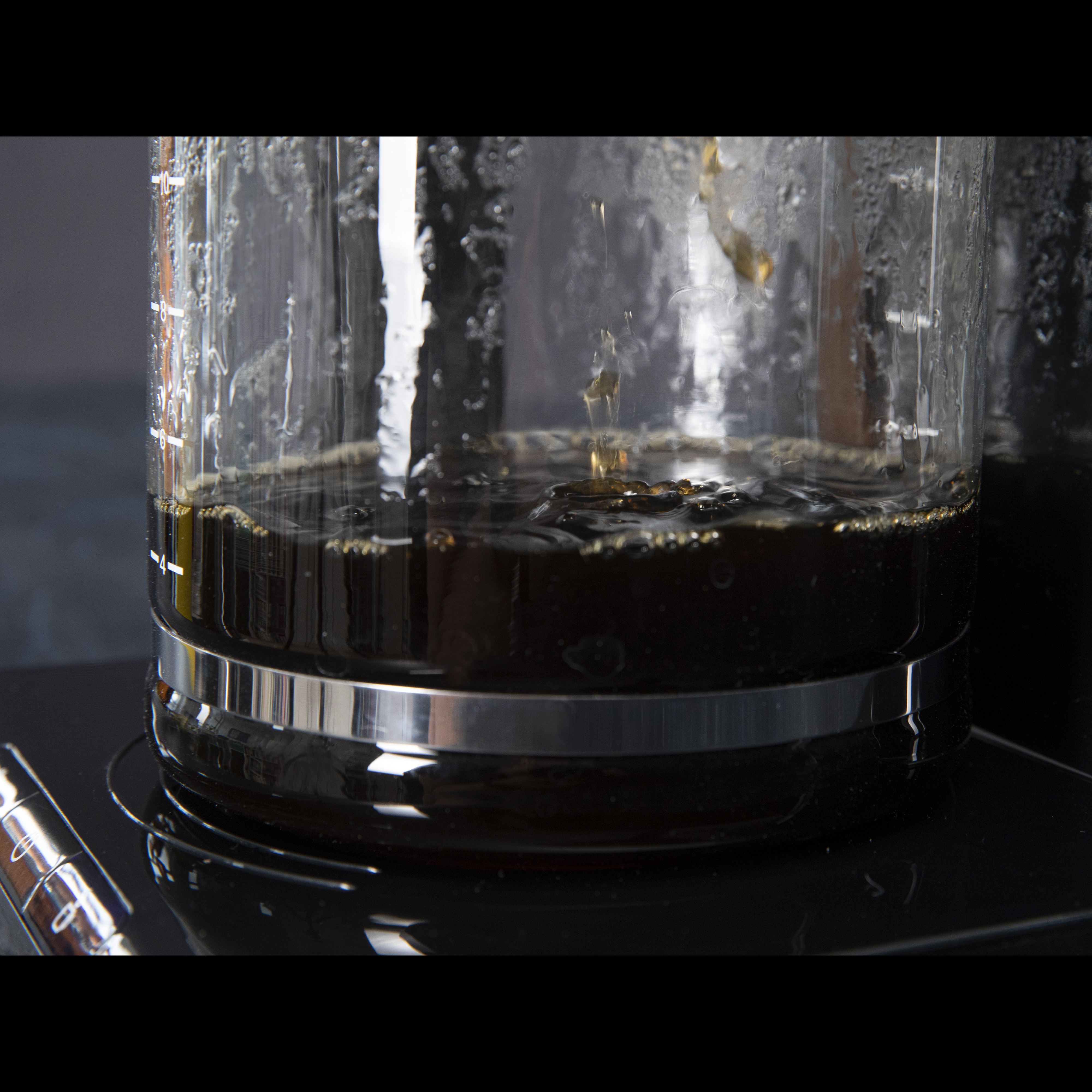Clear glass coffee maker with dark liquid inside, drops falling. Black base, dark background.