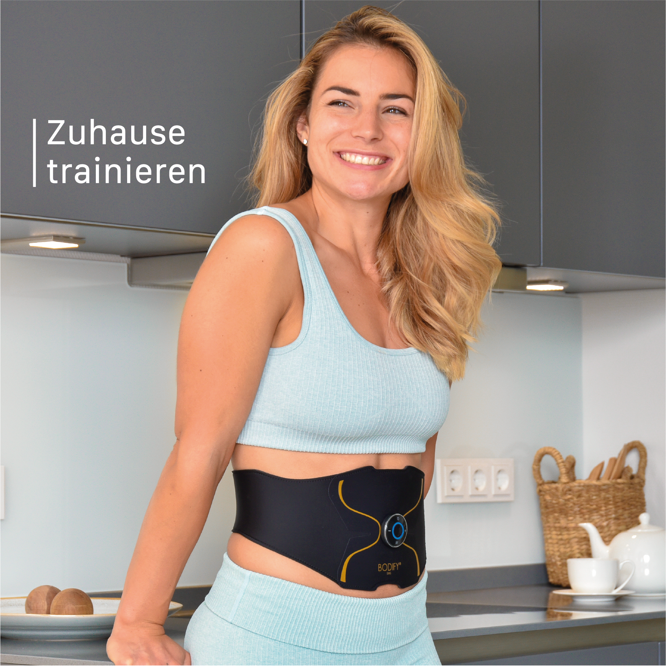 Frau trägt Workout-Gürtel in der Küche. Text lautet 'Zuhause trainieren'. Sie lächelt und schaut nach rechts.