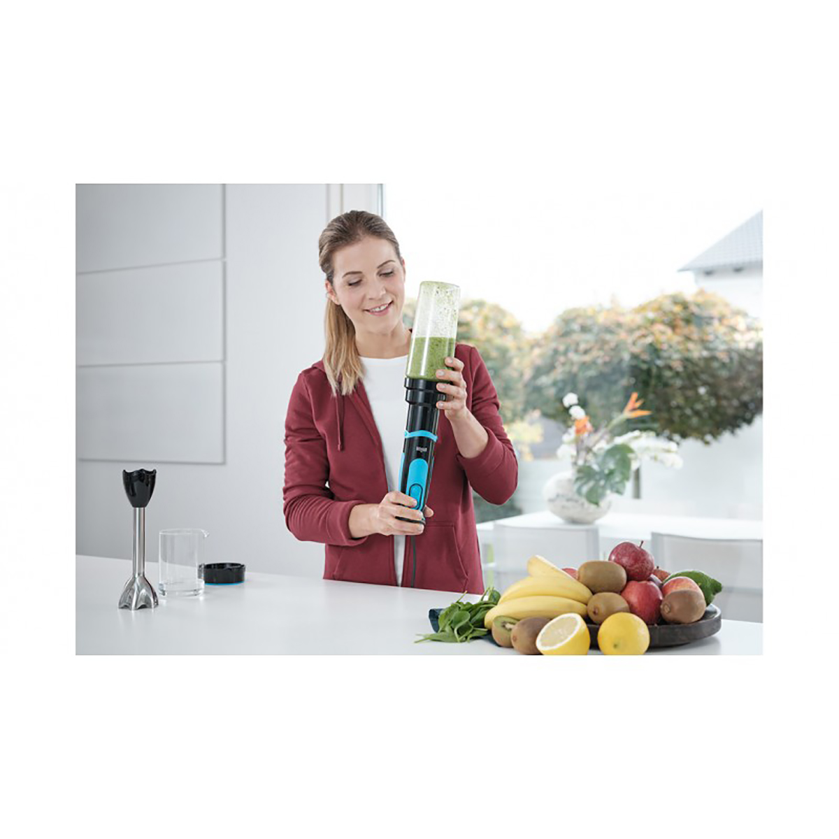 Kadın yeşil smoothie ile blender tutuyor, masada meyveler.