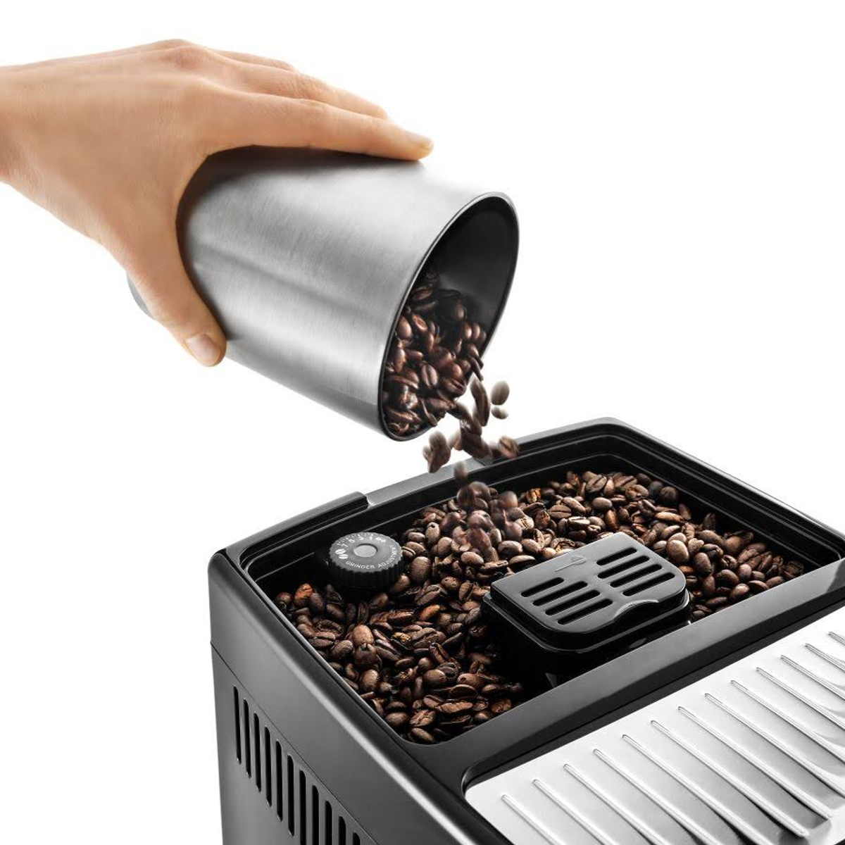 Mano vertiendo granos de café desde un recipiente plateado en la tolva de granos de una cafetera.