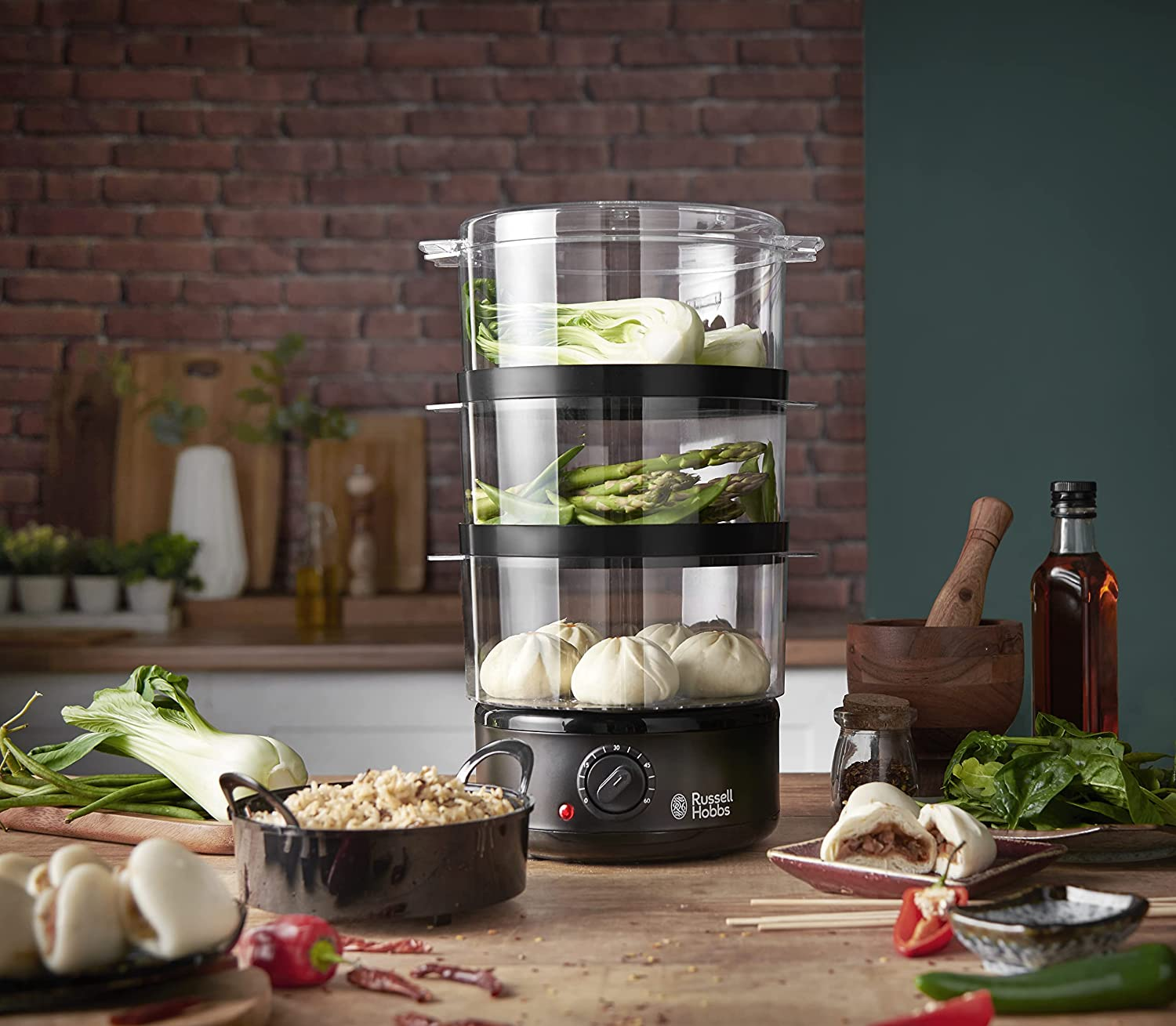 Vaporera con capas de comida: bok choy, espárragos y bollos, con guarniciones en una mesa.