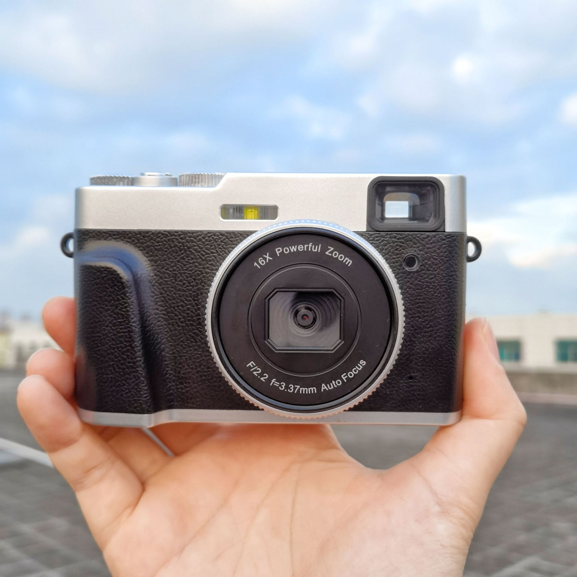 Aparat trzymany w dłoni. Aparat jest czarny i srebrny. Posiada obiektyw z zoomem.