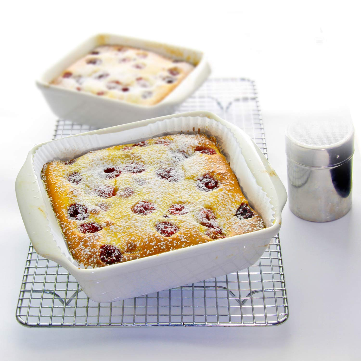 Zwei quadratische Kirsch-Clafoutis in weißen Backformen, mit Puderzucker bestäubt. Gitterrost und Shaker in der Nähe.