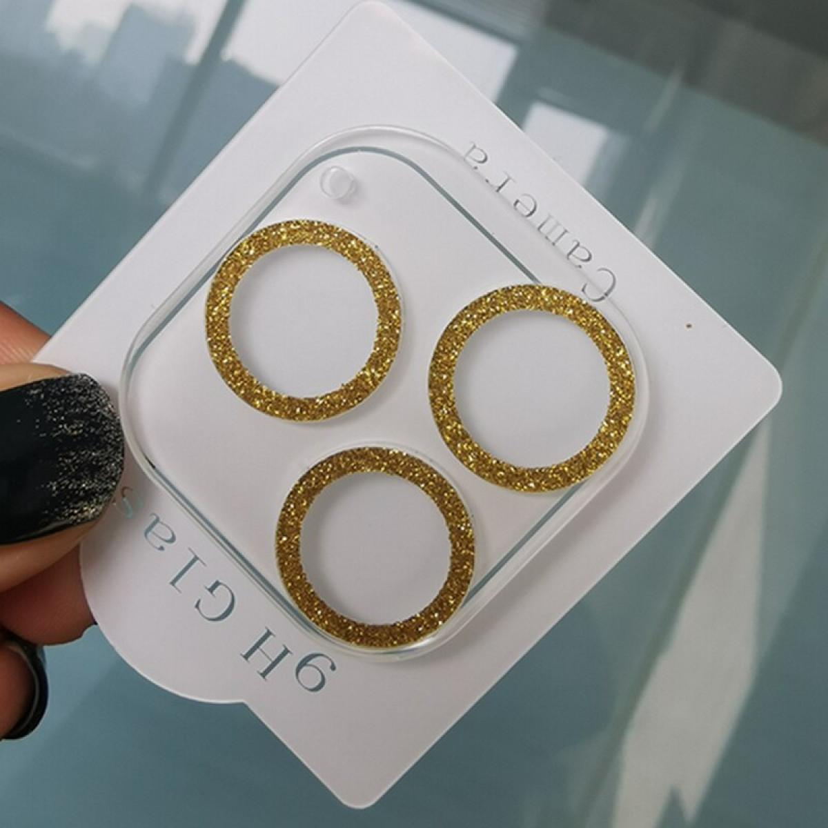 Protezione obiettivo fotocamera del telefono con tre anelli dorati su sfondo bianco con un'unghia nera.