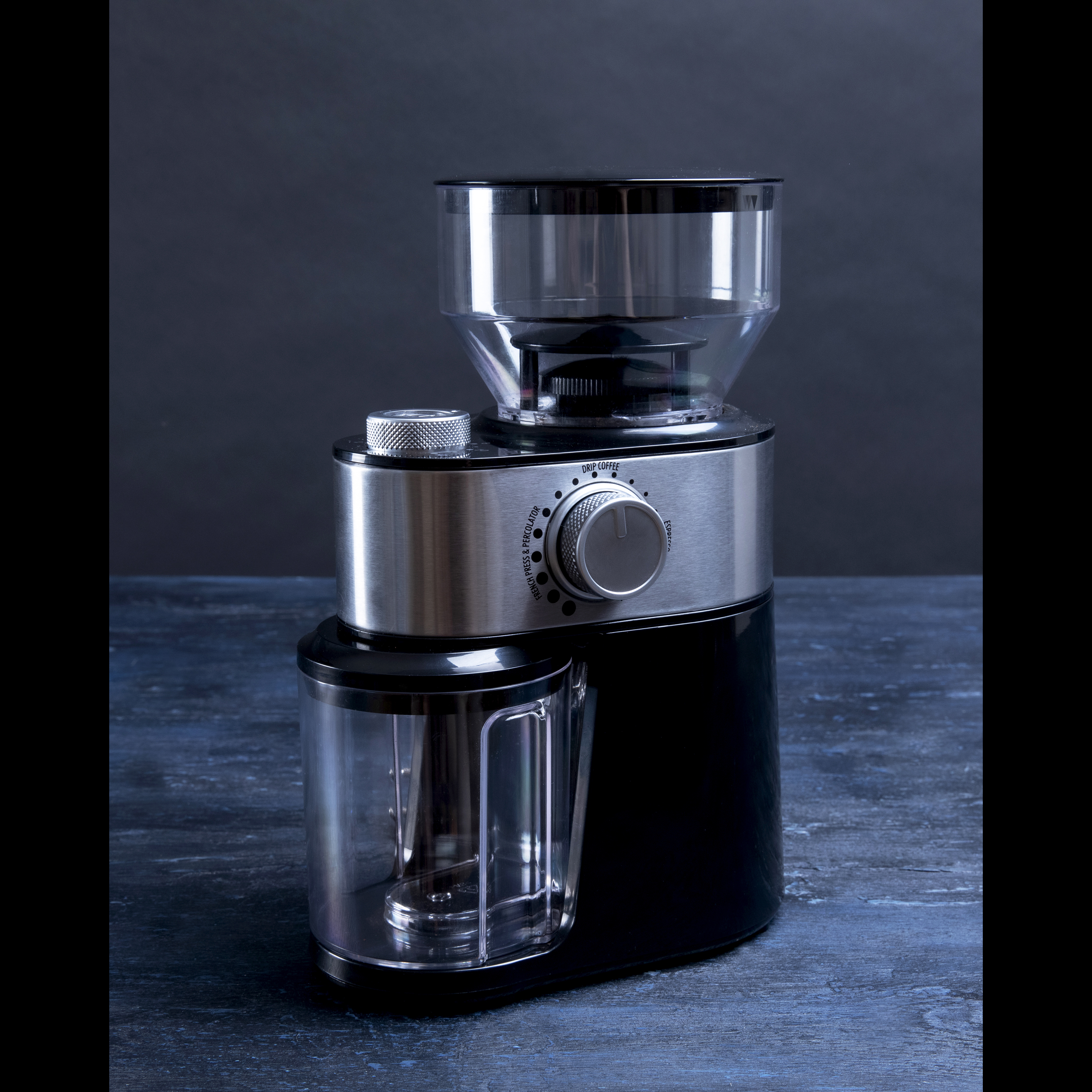 Młynek do kawy: srebrny i czarny z przezroczystym pojemnikiem, na niebieskim drewnianym stole.