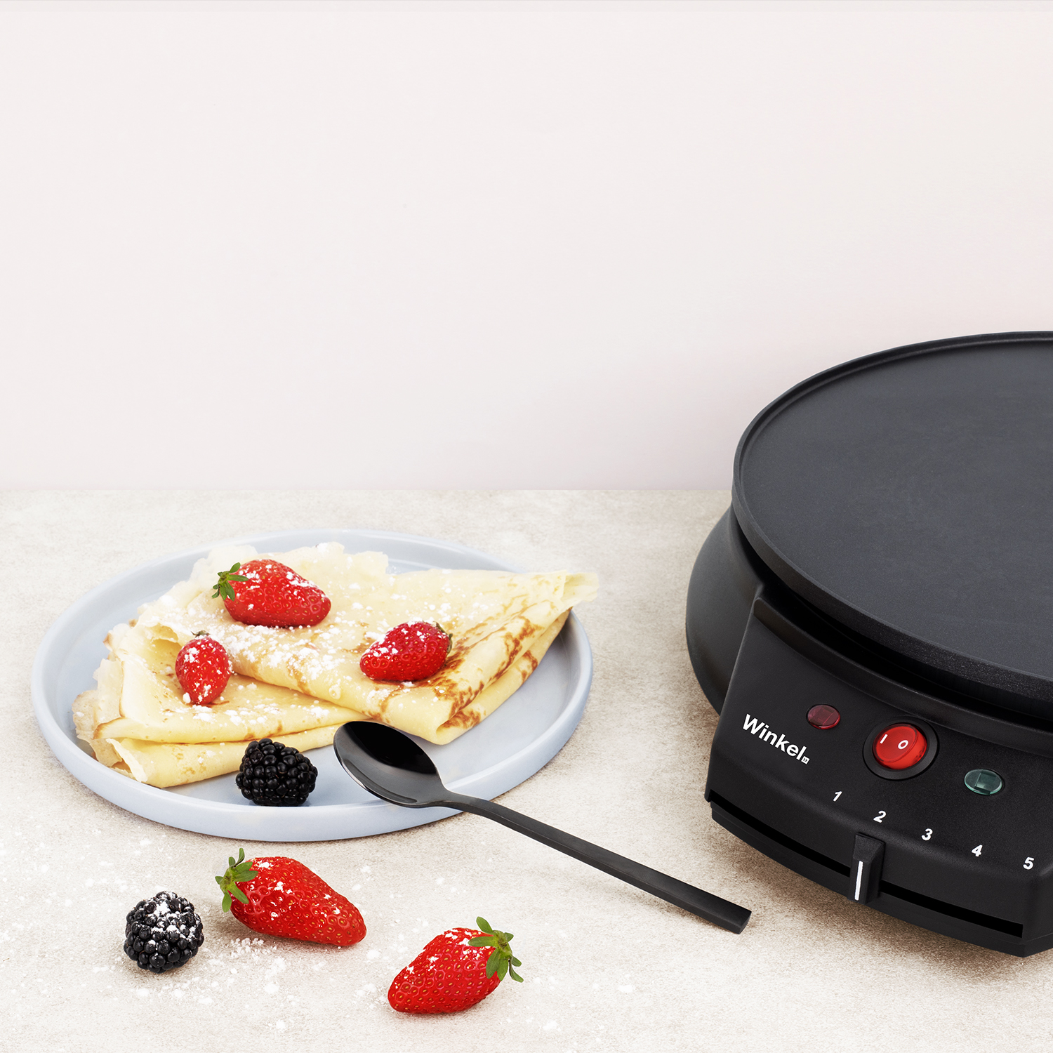 Ein Crêpes-Maker mit Pfannkuchen auf einem Teller, garniert mit Erdbeeren und Brombeeren, ein Löffel daneben.