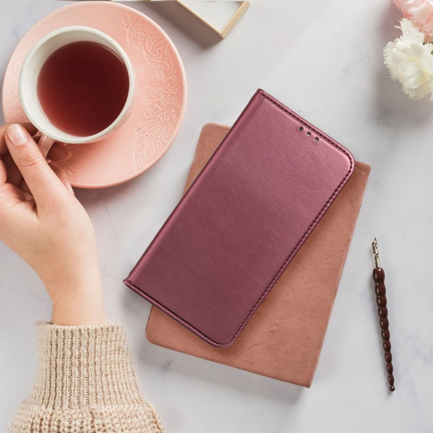 Draufsicht mit einer Hand, die eine Teetasse, Handyhülle, Stift und Blumen hält.