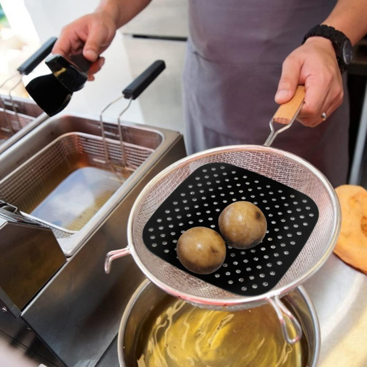 Eine Person brät Essen. Auf einem Sieb befinden sich zwei Gegenstände in einer Fritteuse. In der linken Hand eine Bürste.