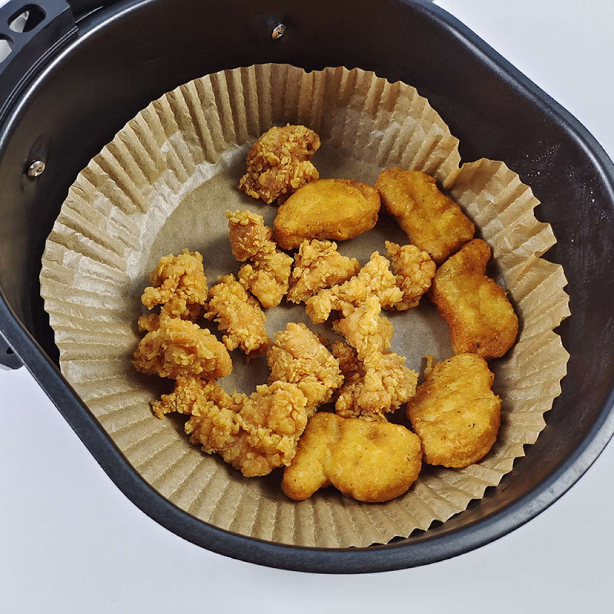 Heißluftfritteuse mit gebratenen Chicken Nuggets und Stücken darin. Das Essen befindet sich auf einer Papiereinlage.