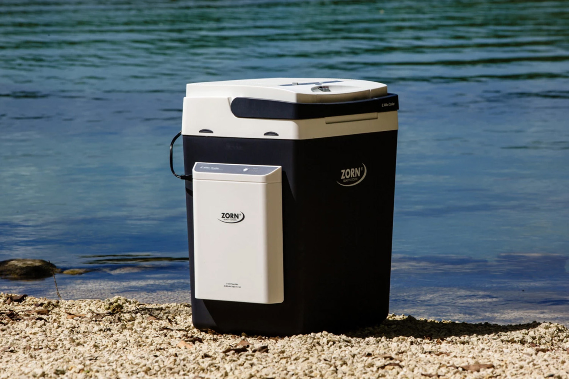 Kühler mit weißem Deckel und schwarzem Körper, am Strand mit Wasser im Hintergrund.
