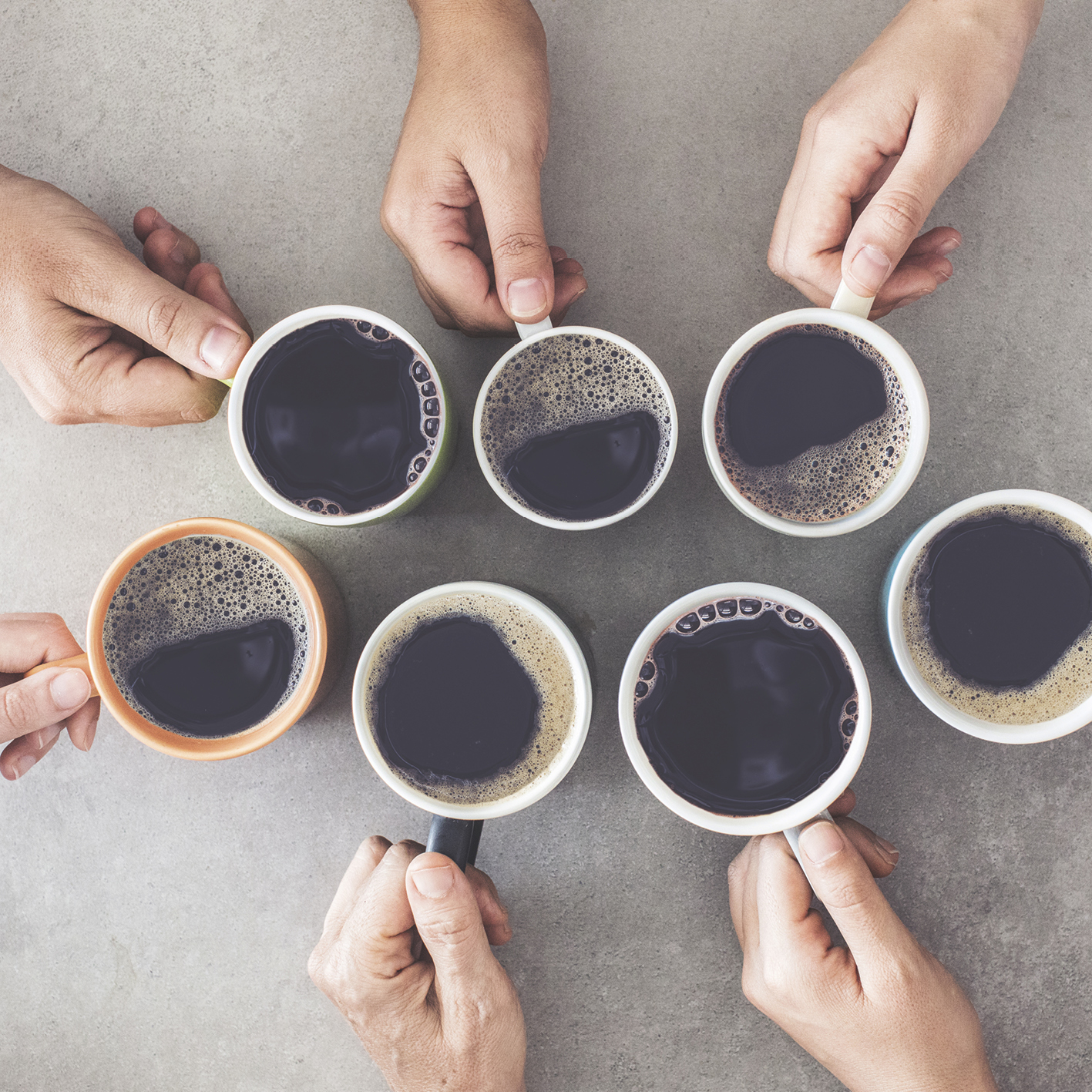 Dłonie trzymające filiżanki z kawą, widok z góry na szarej powierzchni. Różne kolory filiżanek.