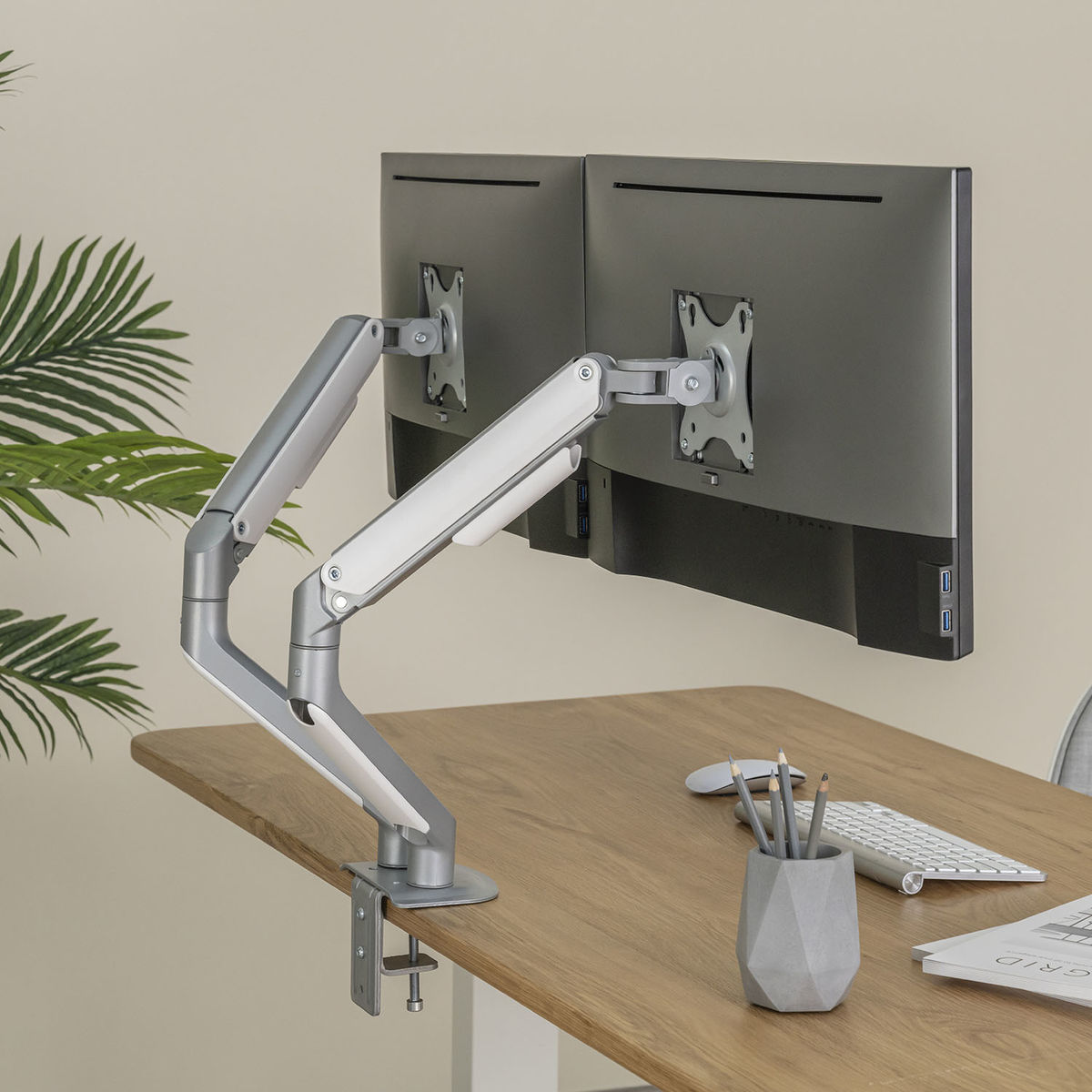 Dubbele monitoropstelling op een bureau met een armbevestiging, potloden en een toetsenbord.