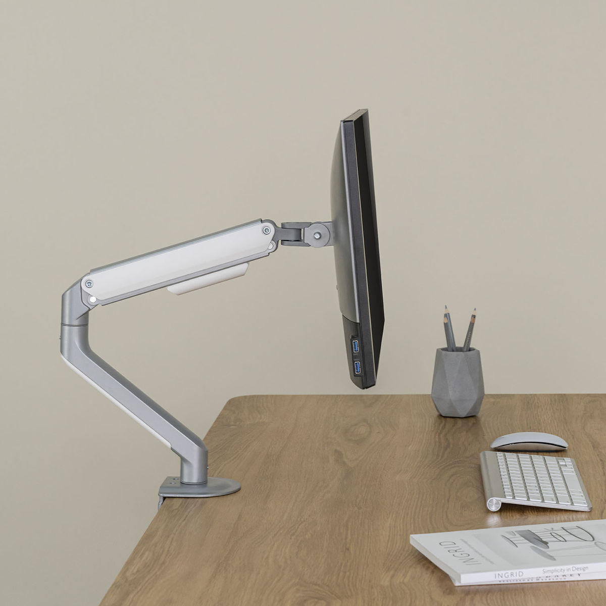 Un moniteur d'ordinateur est monté sur un bras argenté. Le bras est fixé à un bureau en bois. Un porte-crayons est présent.
