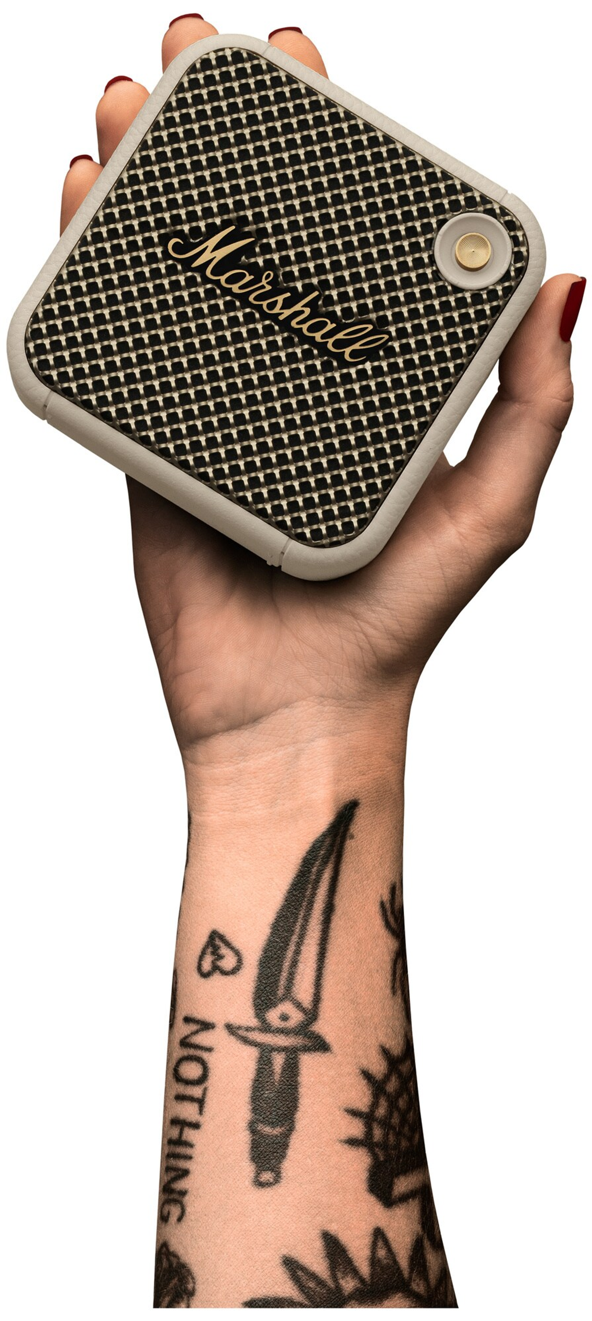 Mano sujetando un altavoz negro con borde crema. Brazo tatuado visible.