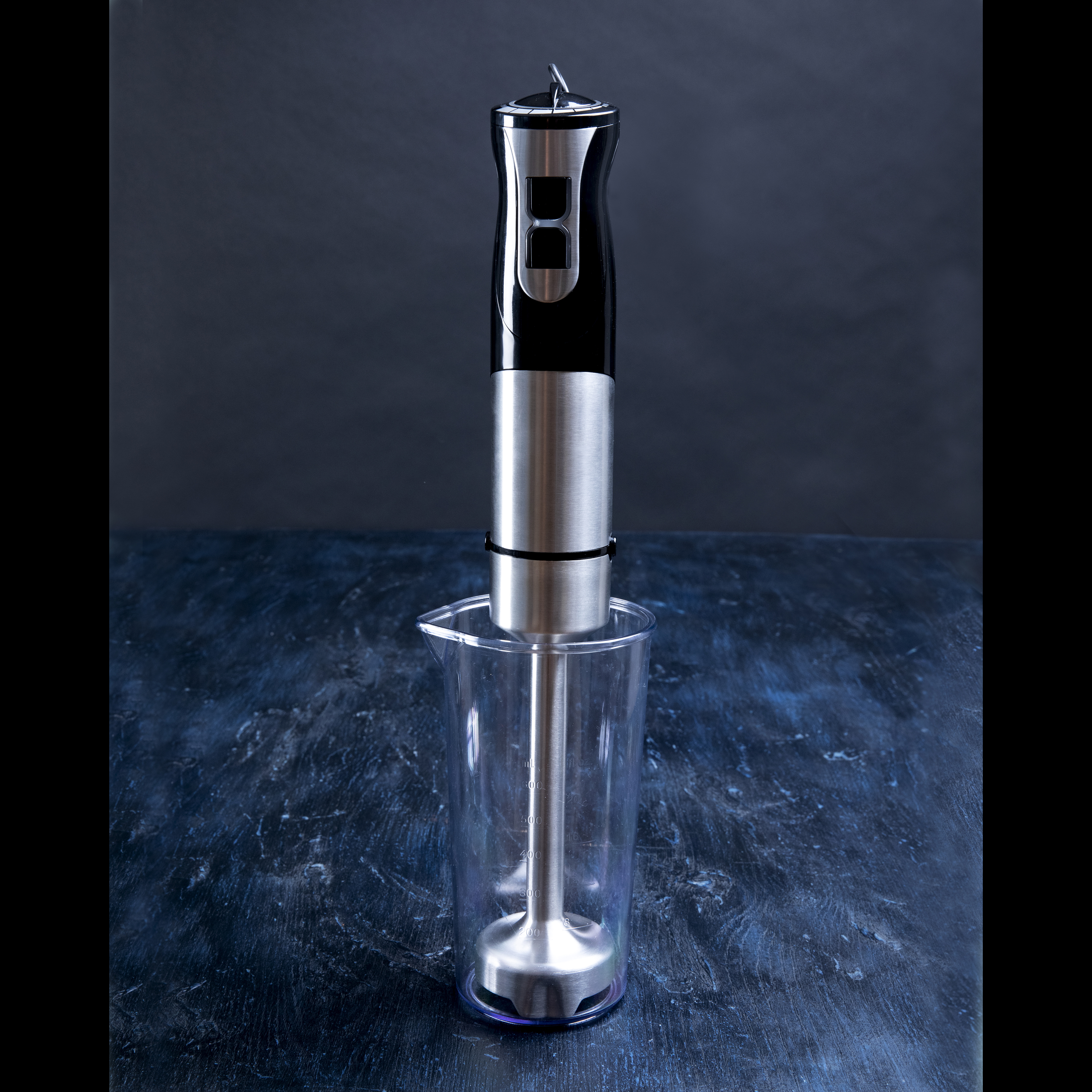 Blender ręczny w przezroczystym kubku na ciemnej powierzchni. Blender jest srebrny i czarny.