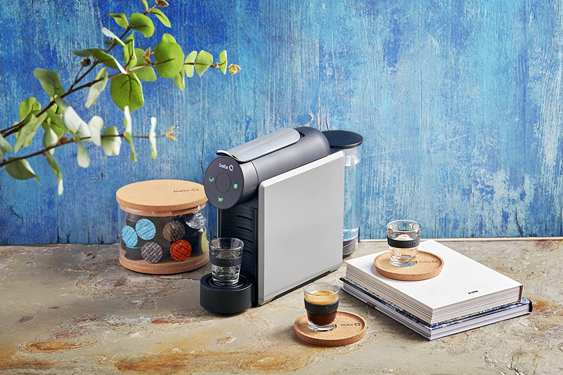Macchina da caffè con tazze e cialde, su tavolo bianco con sfondo blu.
