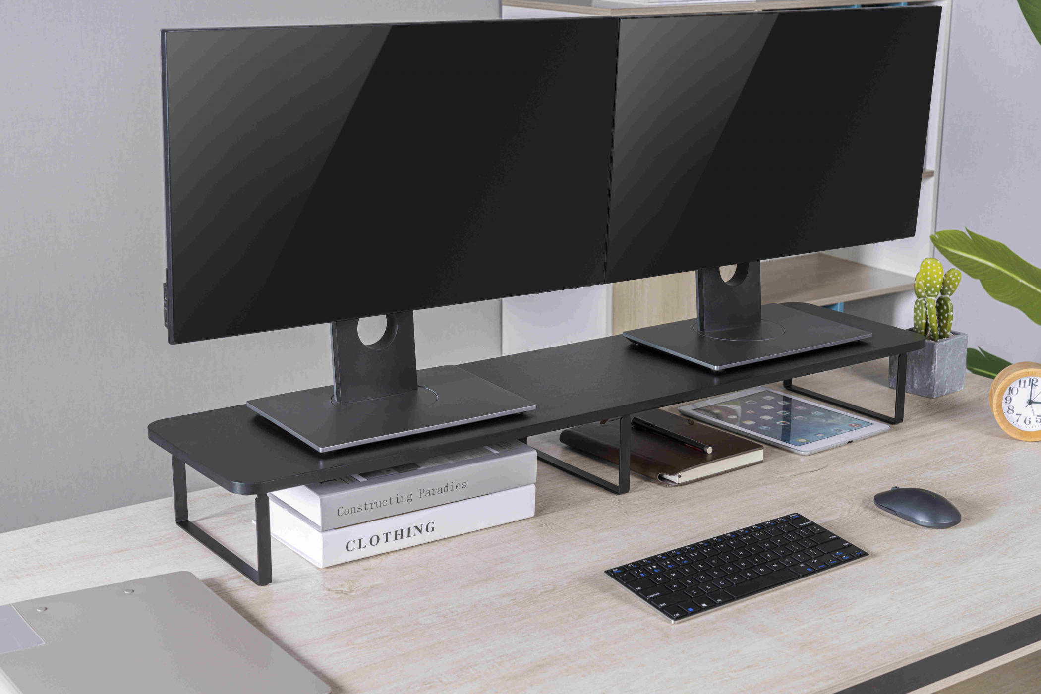 Zwei Monitore auf einem schwarzen Ständer, mit Tastatur, Tablet und Büchern auf einem Schreibtisch.
