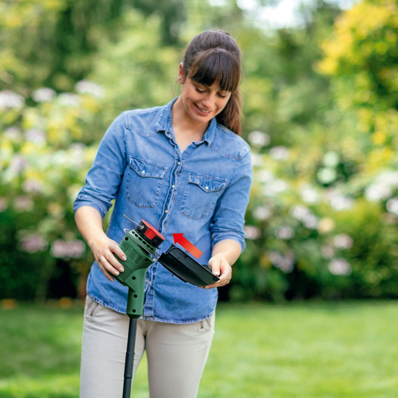 Femme en chemise en jean utilise une tondeuse verte dans jardin herbeux avec arbres.