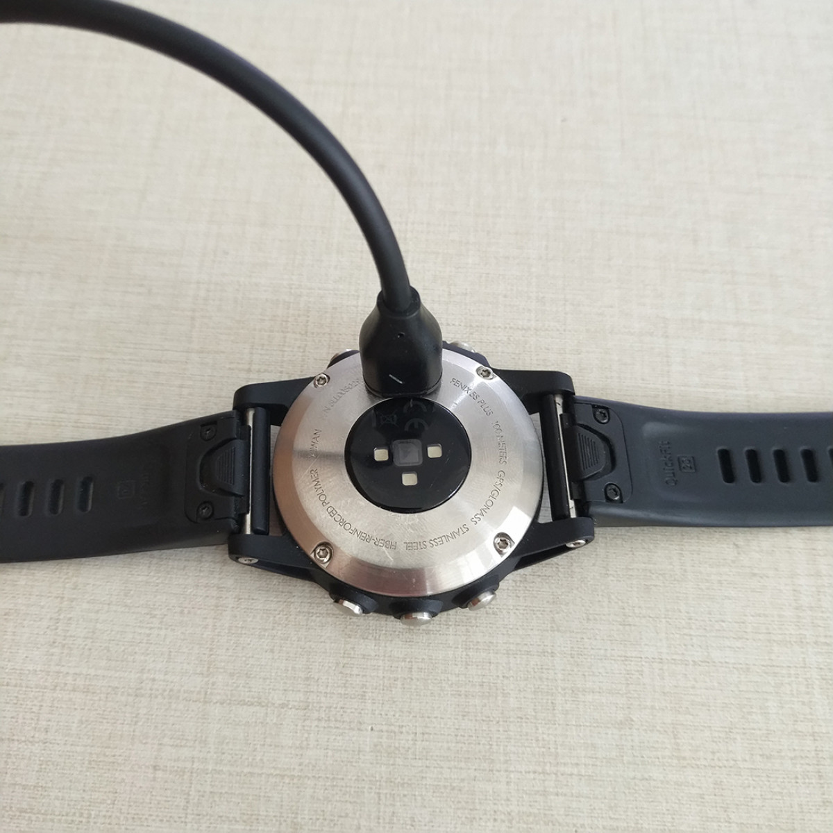 Achterkant smartwatch met oplaadkabel aangesloten. Horloge is zwart, kabel is zwart, op een licht oppervlak.