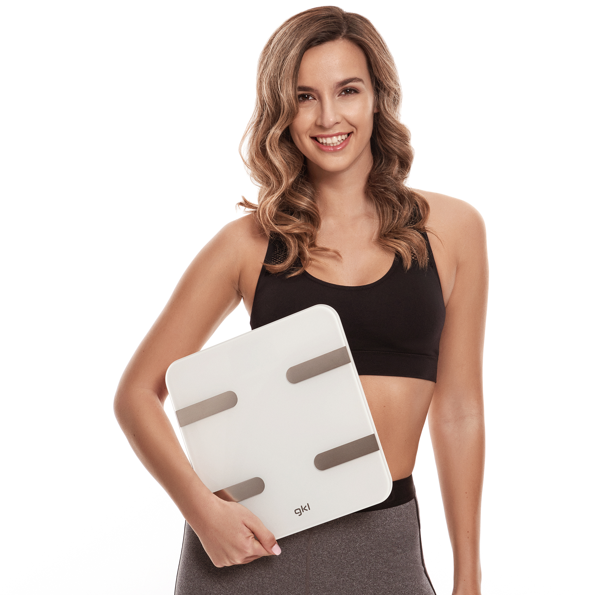 Una mujer sonriente sosteniendo una báscula digital blanca. Lleva un top negro y leggings grises.