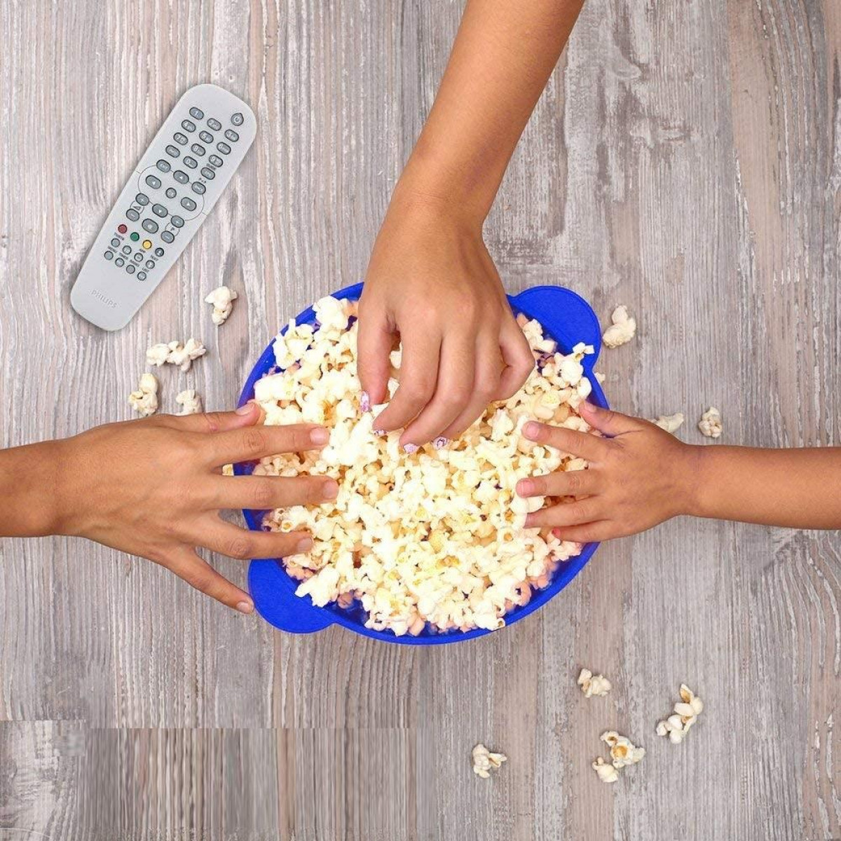 Hands reaching into a blue bowl filled with popcorn, a remote, and popcorn scattered on wood.