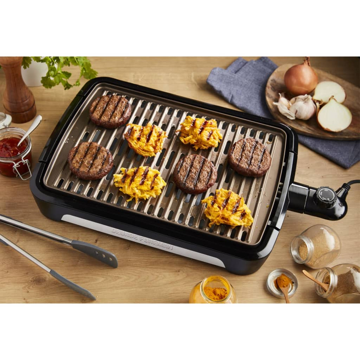Una griglia elettrica con hamburger e verdure cotti su un tavolo di legno con condimenti.