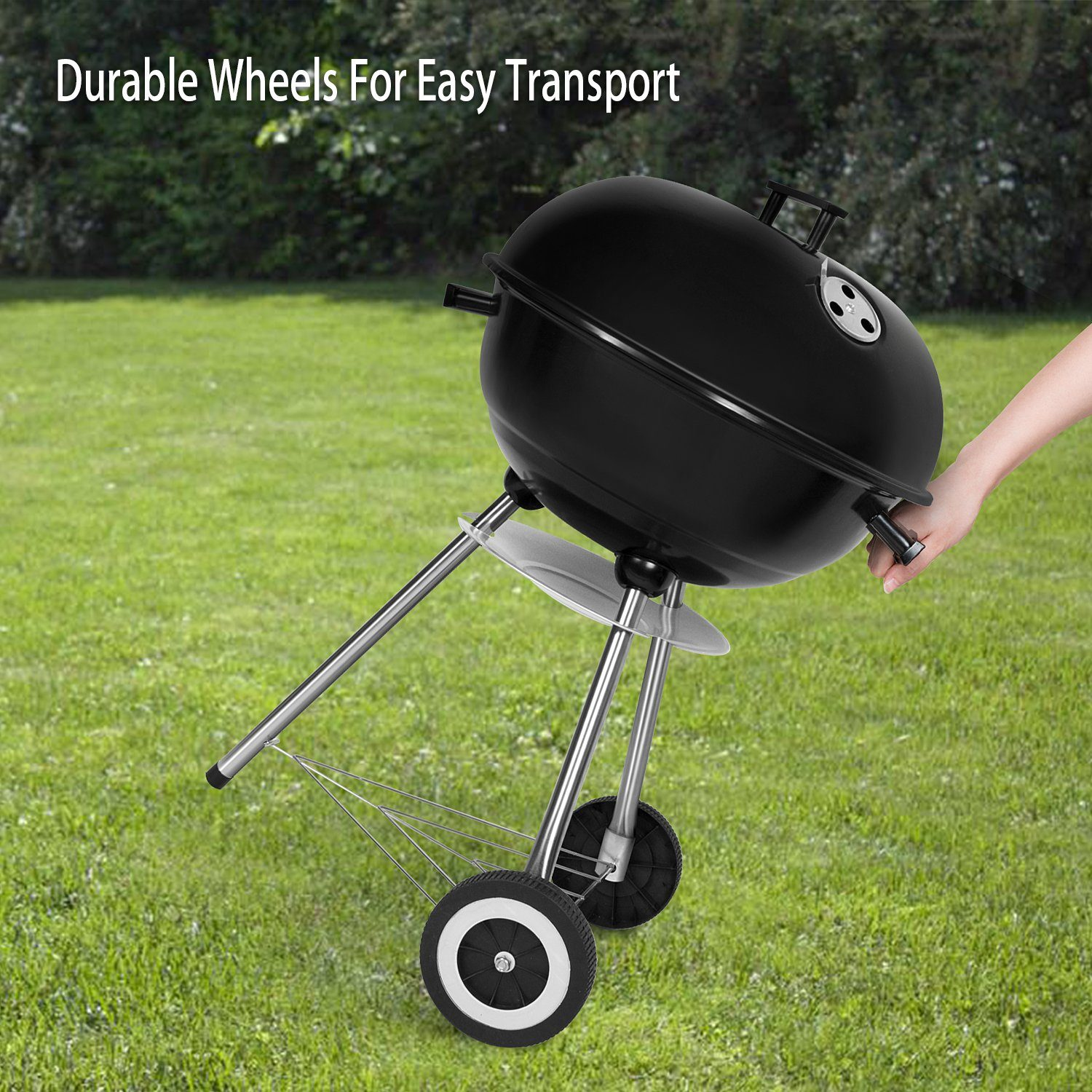 Schwarzer Holzkohlegrill auf grünem Gras, wird von einer Hand am Griff bewegt. Räder sind sichtbar.