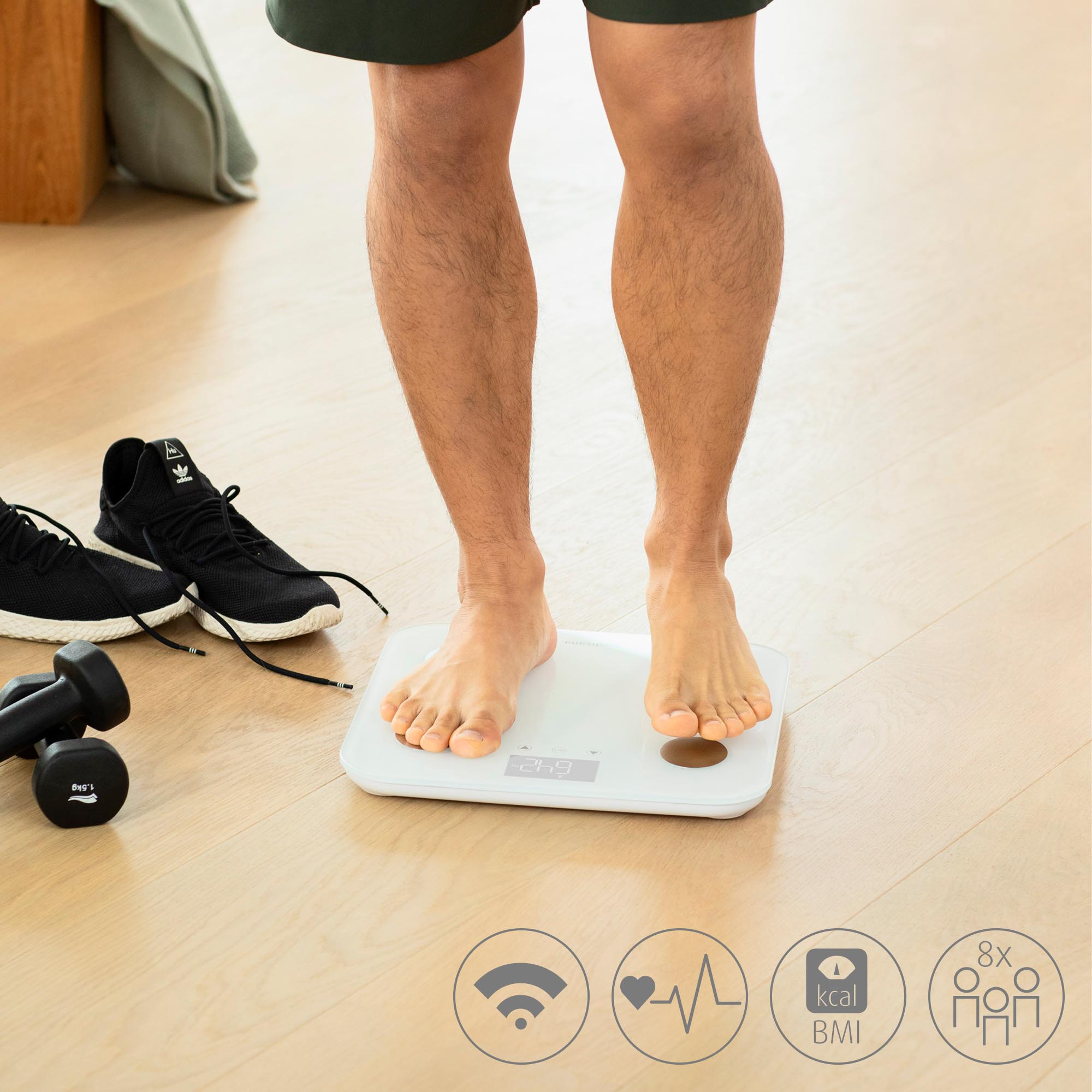 Uomo in piedi su una bilancia digitale bianca. Scarpe da ginnastica nere e pesi sul pavimento.