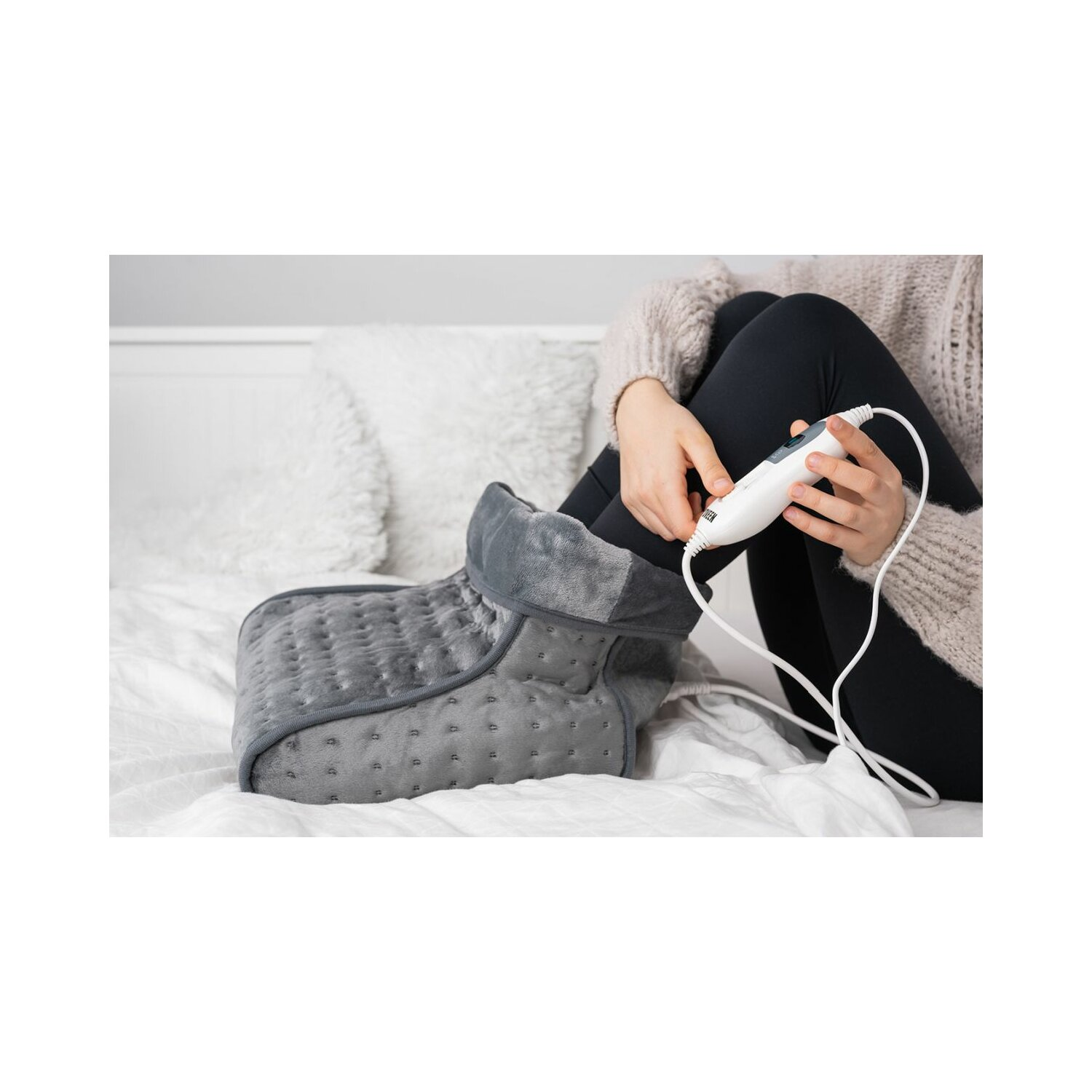 Osoba trzyma szary podgrzewacz do stóp z białym kablem. Osoba siedzi na łóżku.