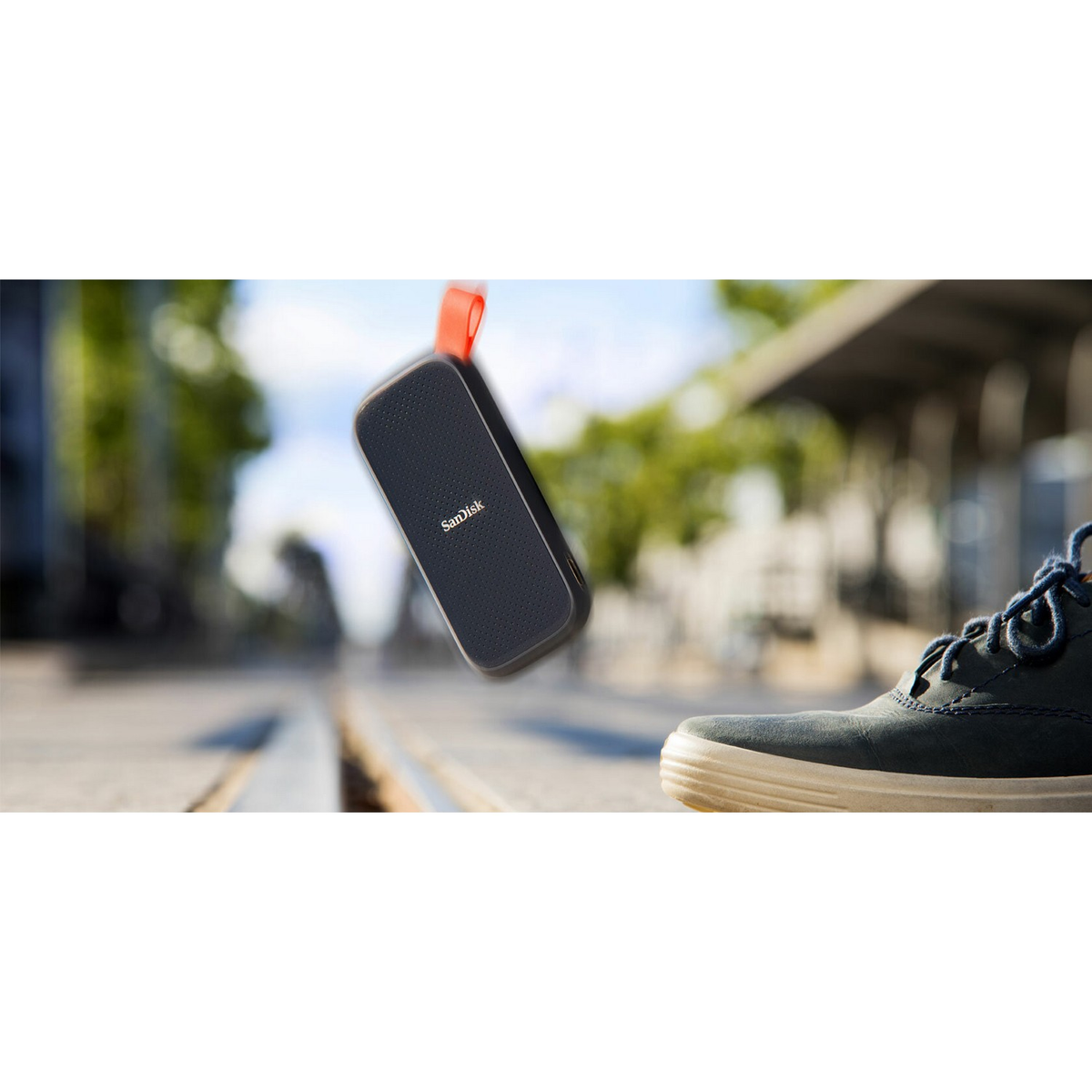 Głośnik SanDisk spada. Czarny, pomarańczowy pasek. Rozmyte tło torów, buty w fokusie.