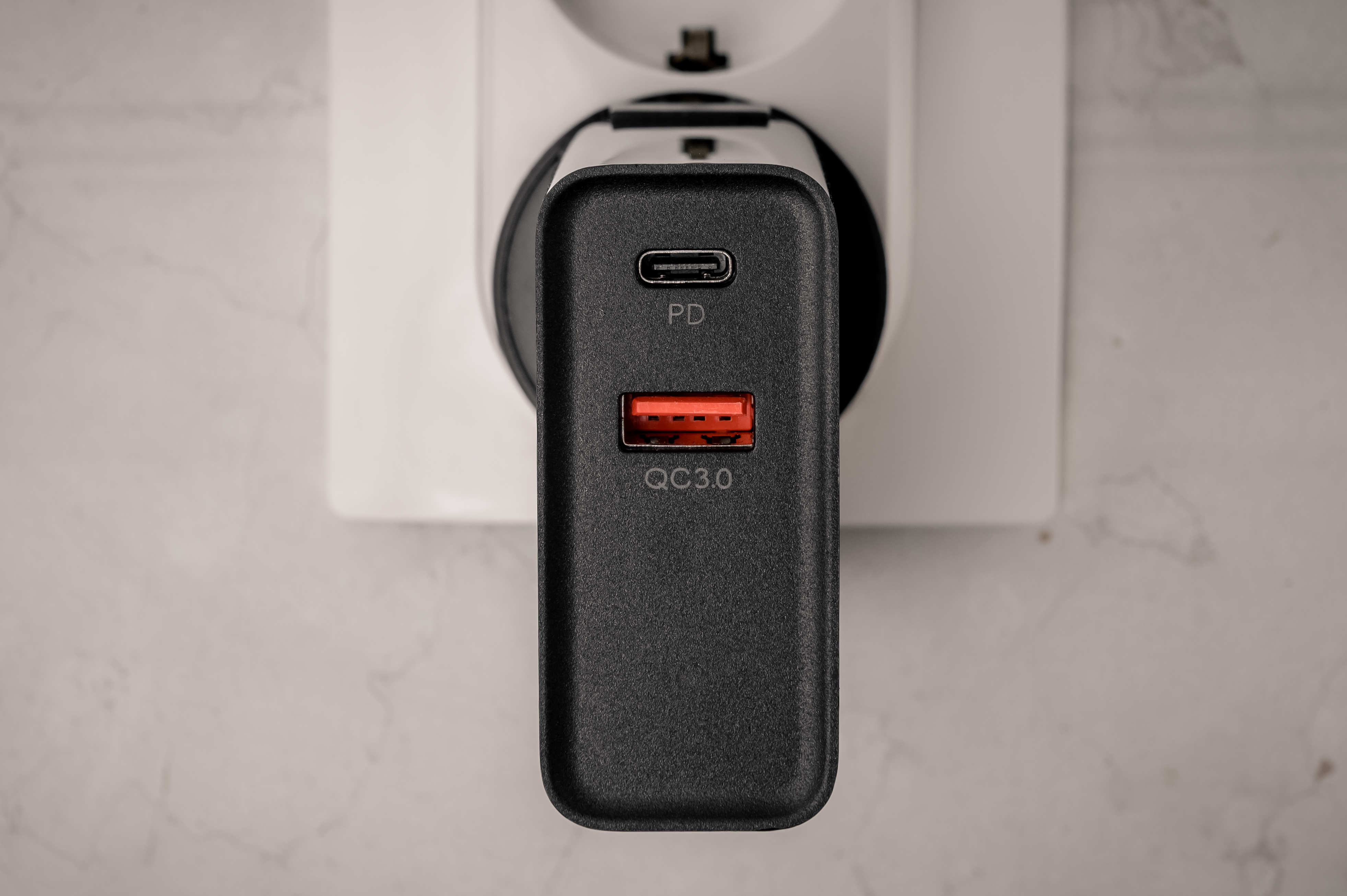 Cargador de pared negro con puertos USB-C y USB-A, enchufado a un enchufe de pared blanco.