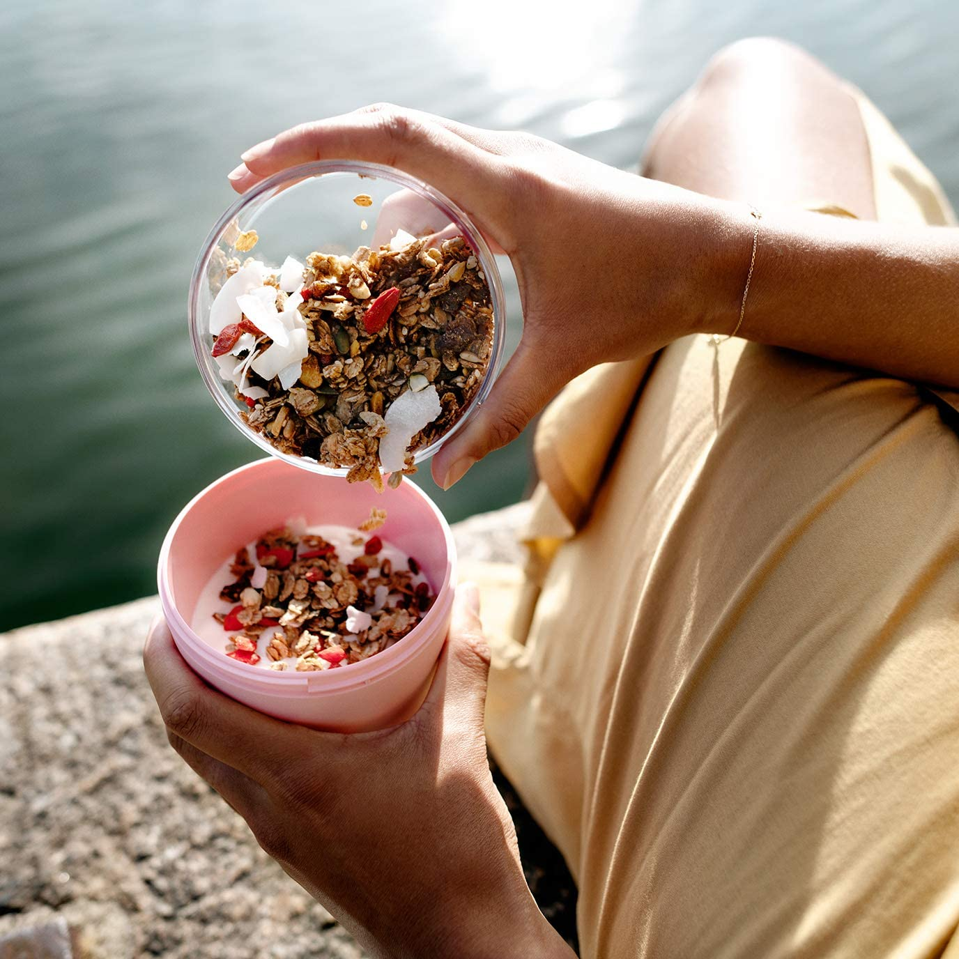 Eine Person gießt Müsli aus einem transparenten Behälter in eine rosa Schüssel mit Joghurt. Im Hintergrund ist ein See.