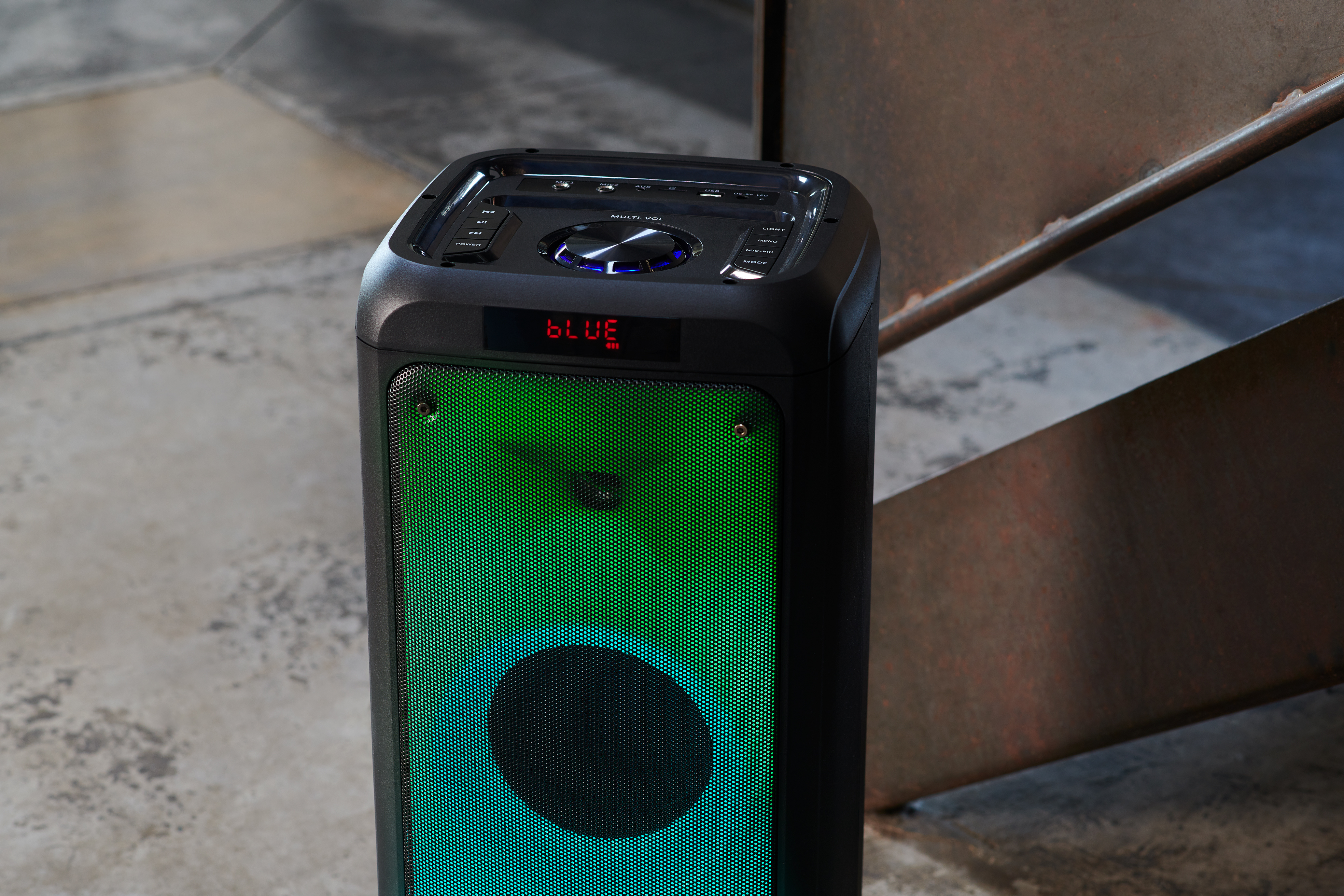 Altavoz negro con luz verde y pantalla azul. Una escalera metálica al fondo.