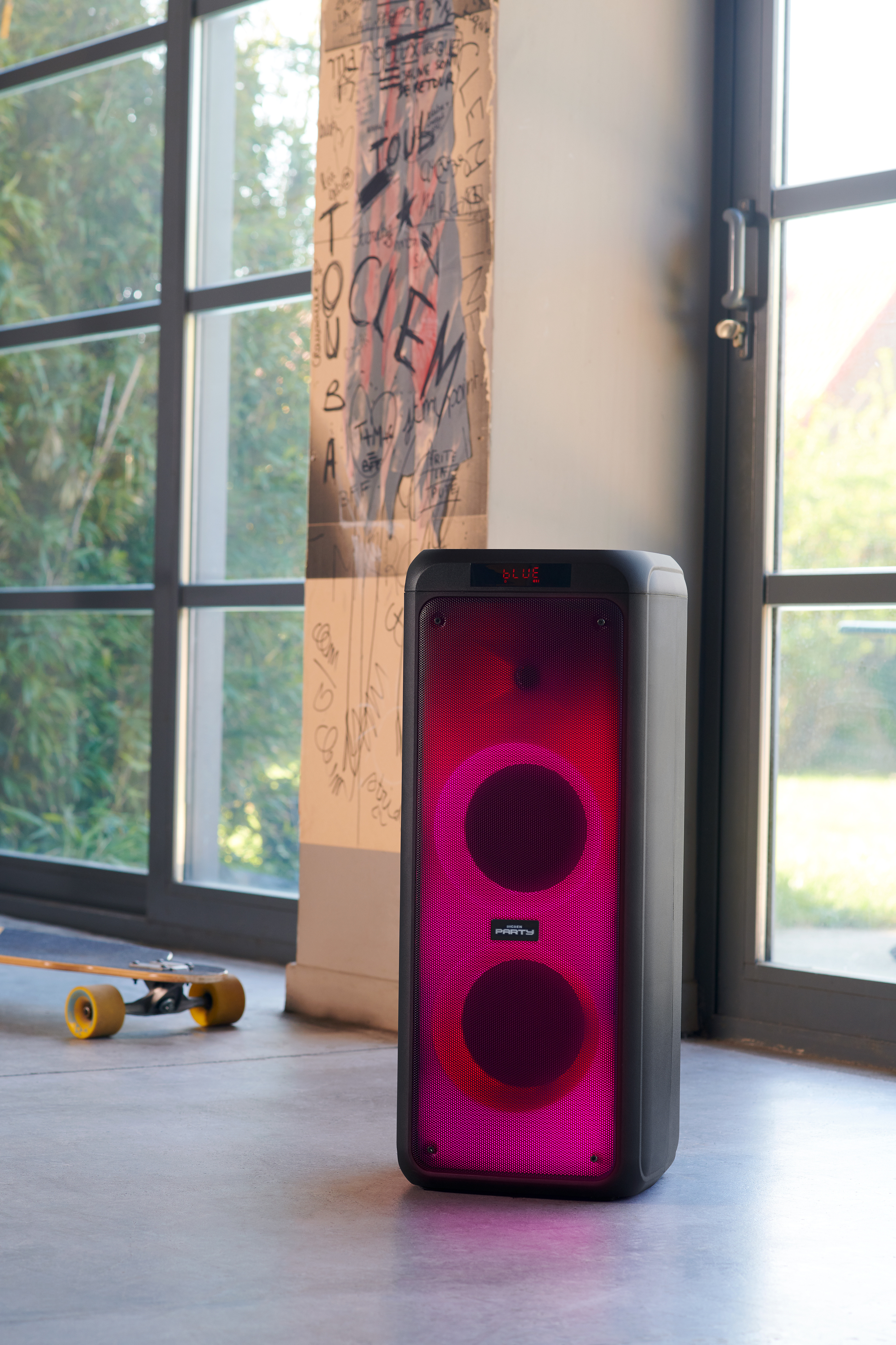 Un altavoz de fiesta negro grande con luces rosas y rojas, junto a una ventana con una patineta.
