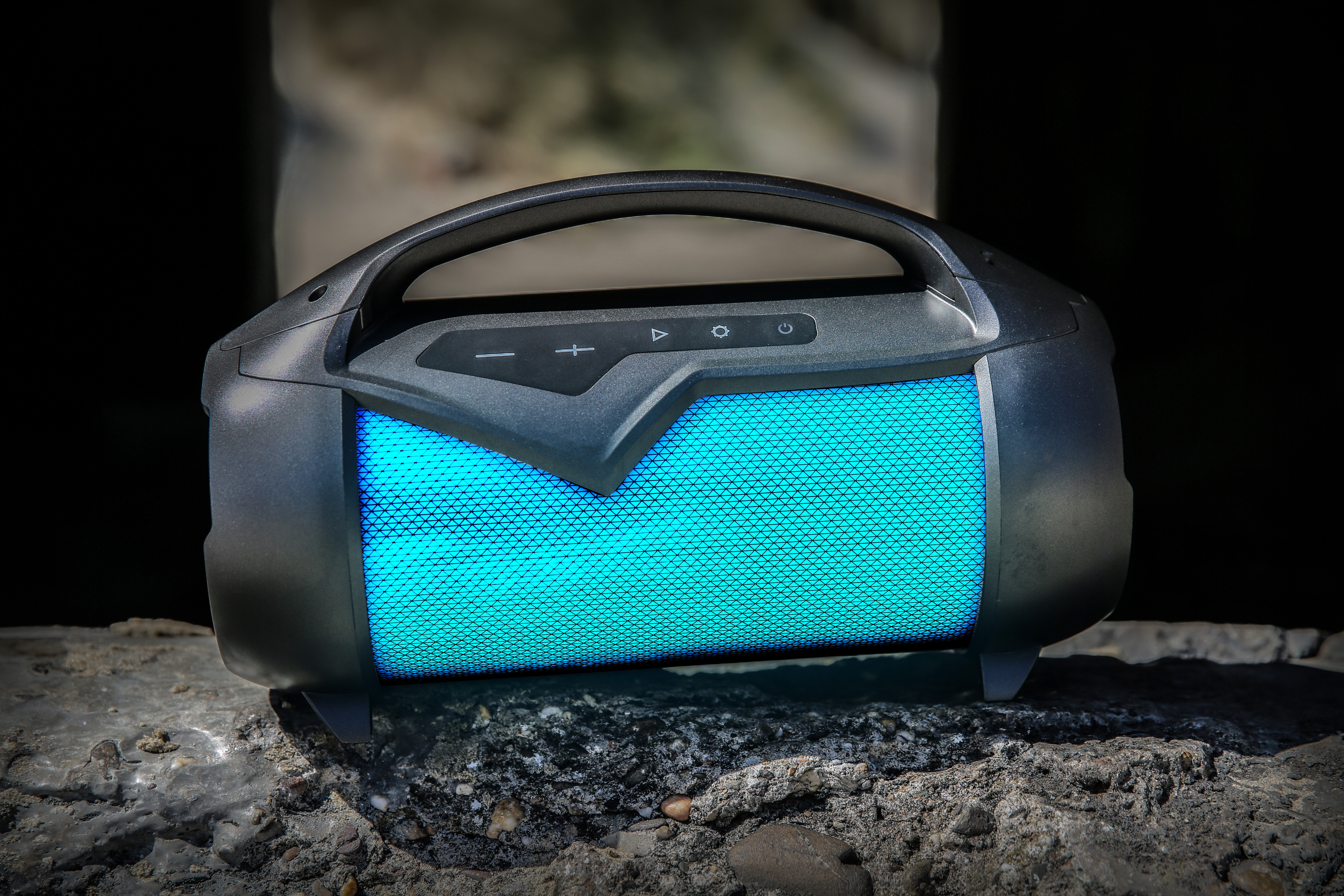 Czarny głośnik z niebieskim światłem, przyciski na górze. Na skalistej powierzchni.