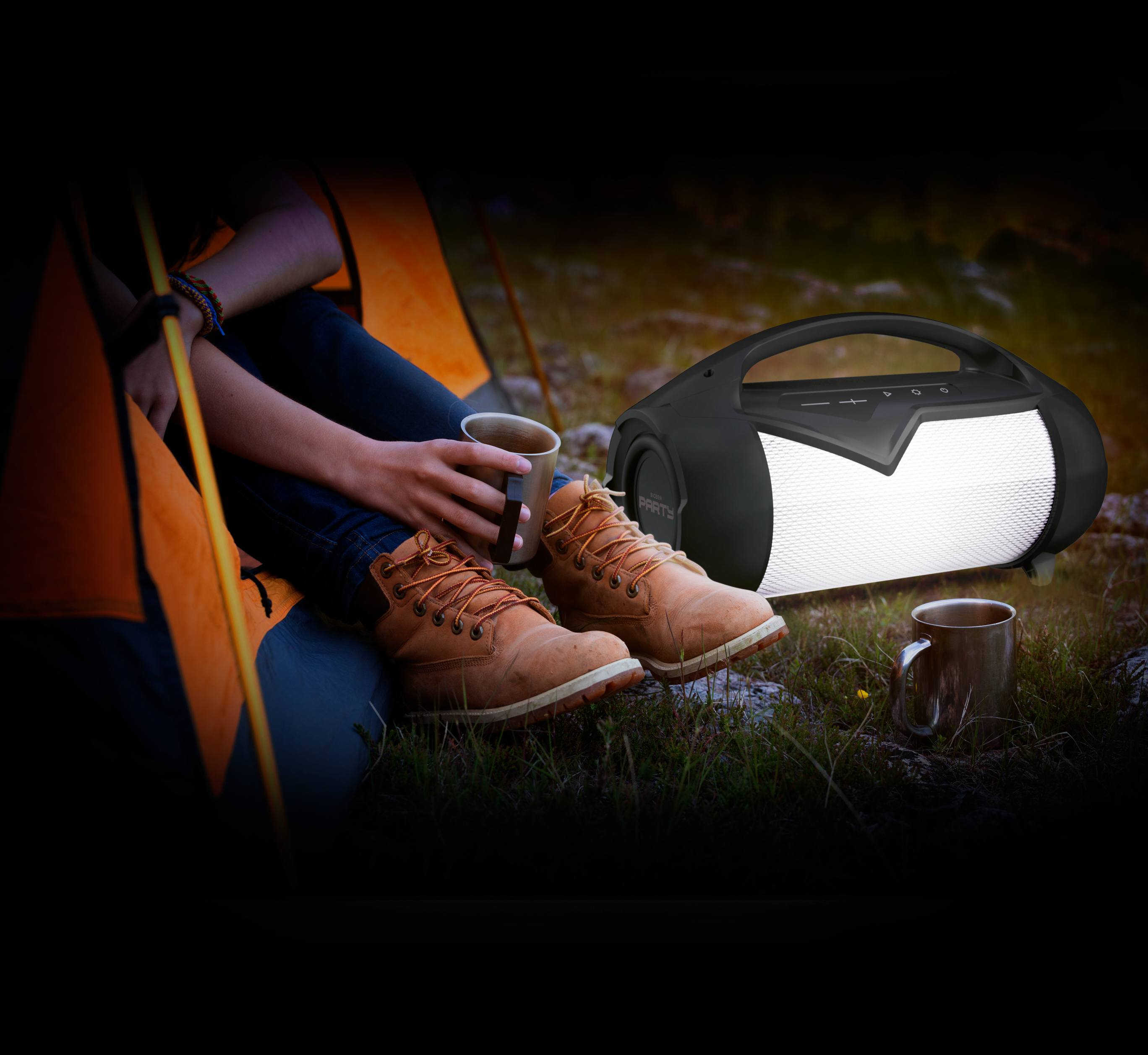 Osoba siedzi z kubkiem i brązowymi butami z głośnikiem po prawej stronie. Namiot w tle.
