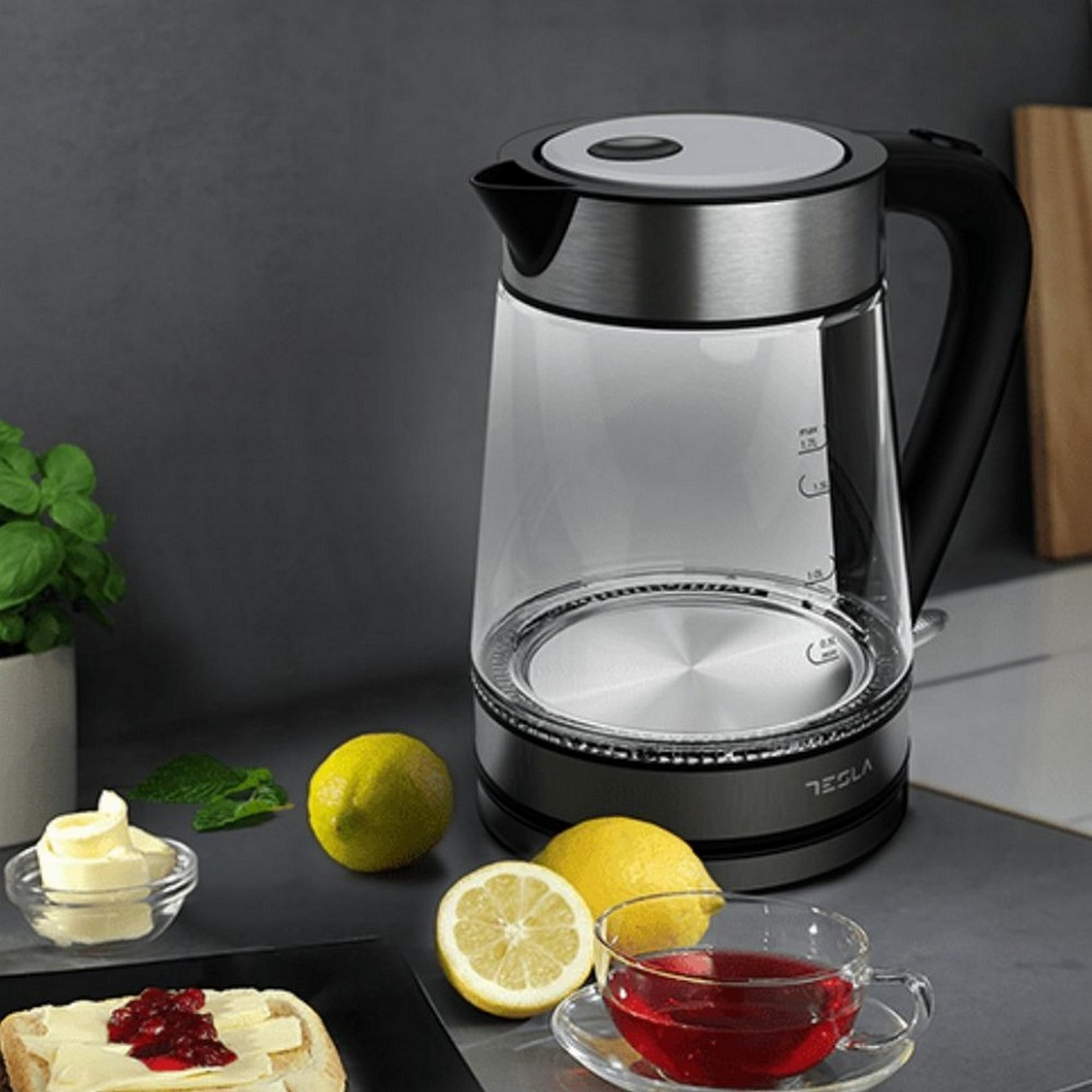 Szklany czajnik elektryczny z herbatą, cytrynami i masłem na szarym blacie.