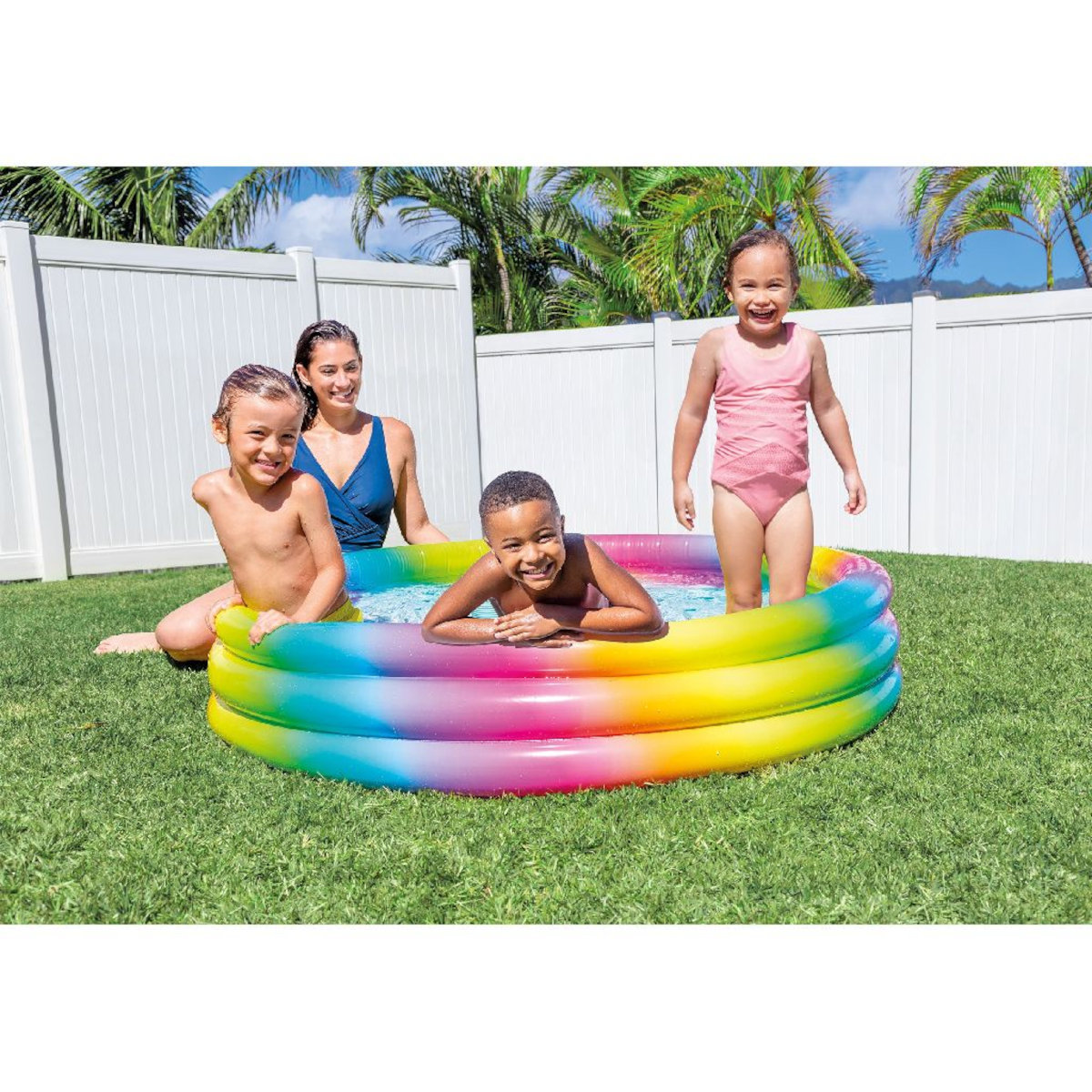 Vier Kinder und eine Frau in einem Regenbogenpool auf grünem Gras. Weißer Zaun und Bäume.