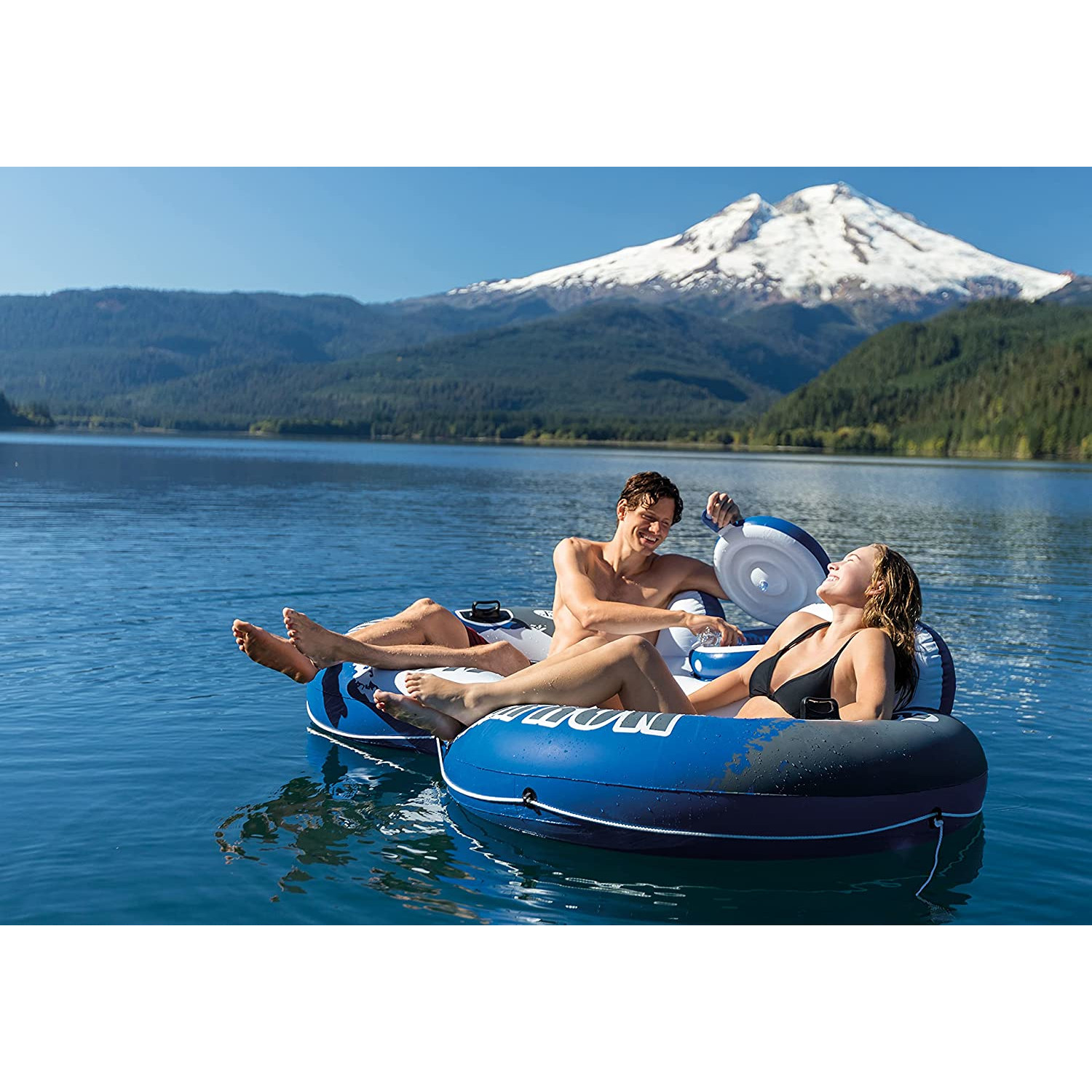 Ein Mann und eine Frau auf aufblasbaren Ringen in einem See, mit einem Berg im Hintergrund.