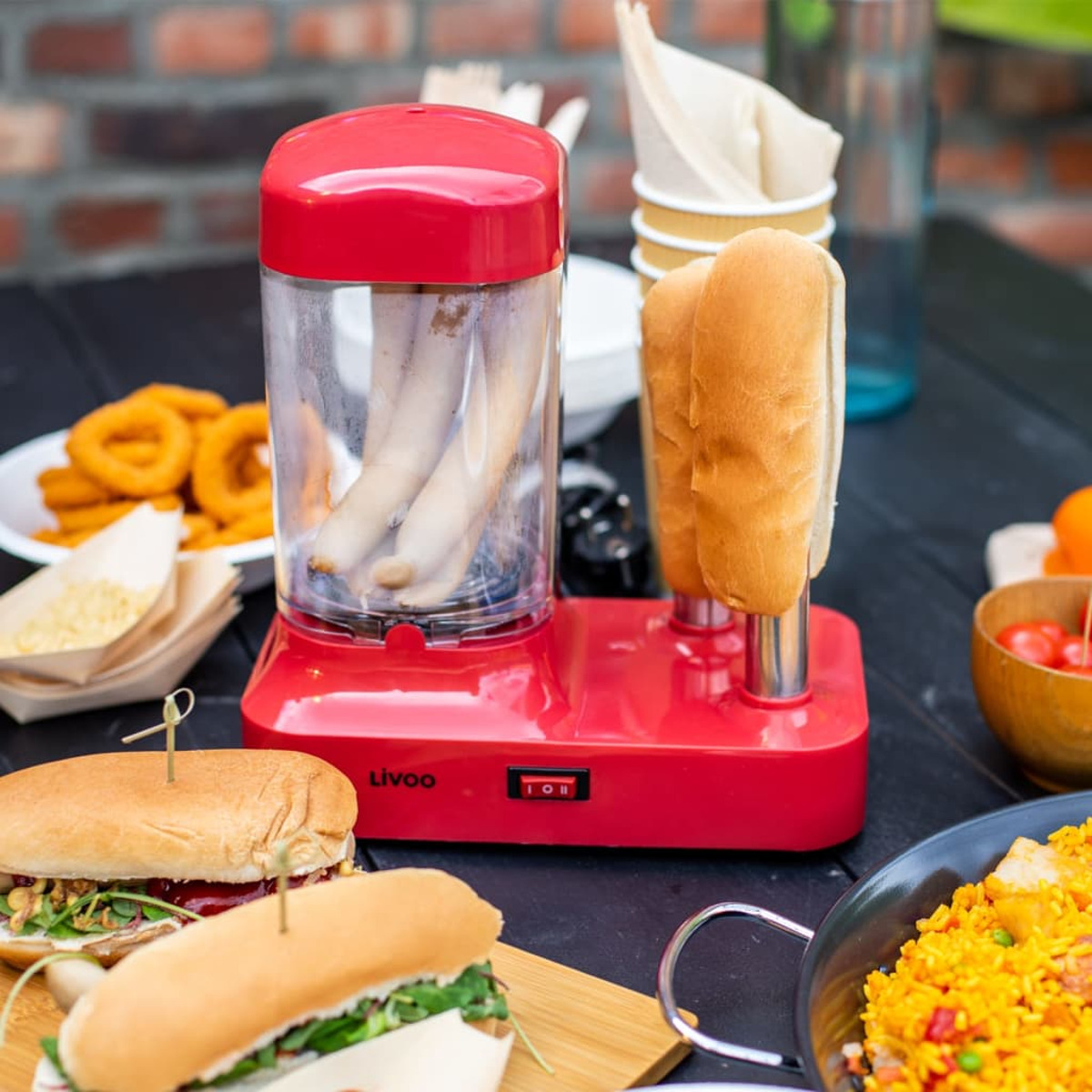 Roter Hotdog-Maker mit zwei Brötchen, andere Lebensmittel und Ziegelhintergrund.