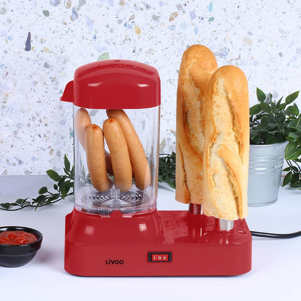 Roter Hotdog-Kocher mit Würstchen und Brot, weiße Arbeitsplatte, grüne Pflanzen.